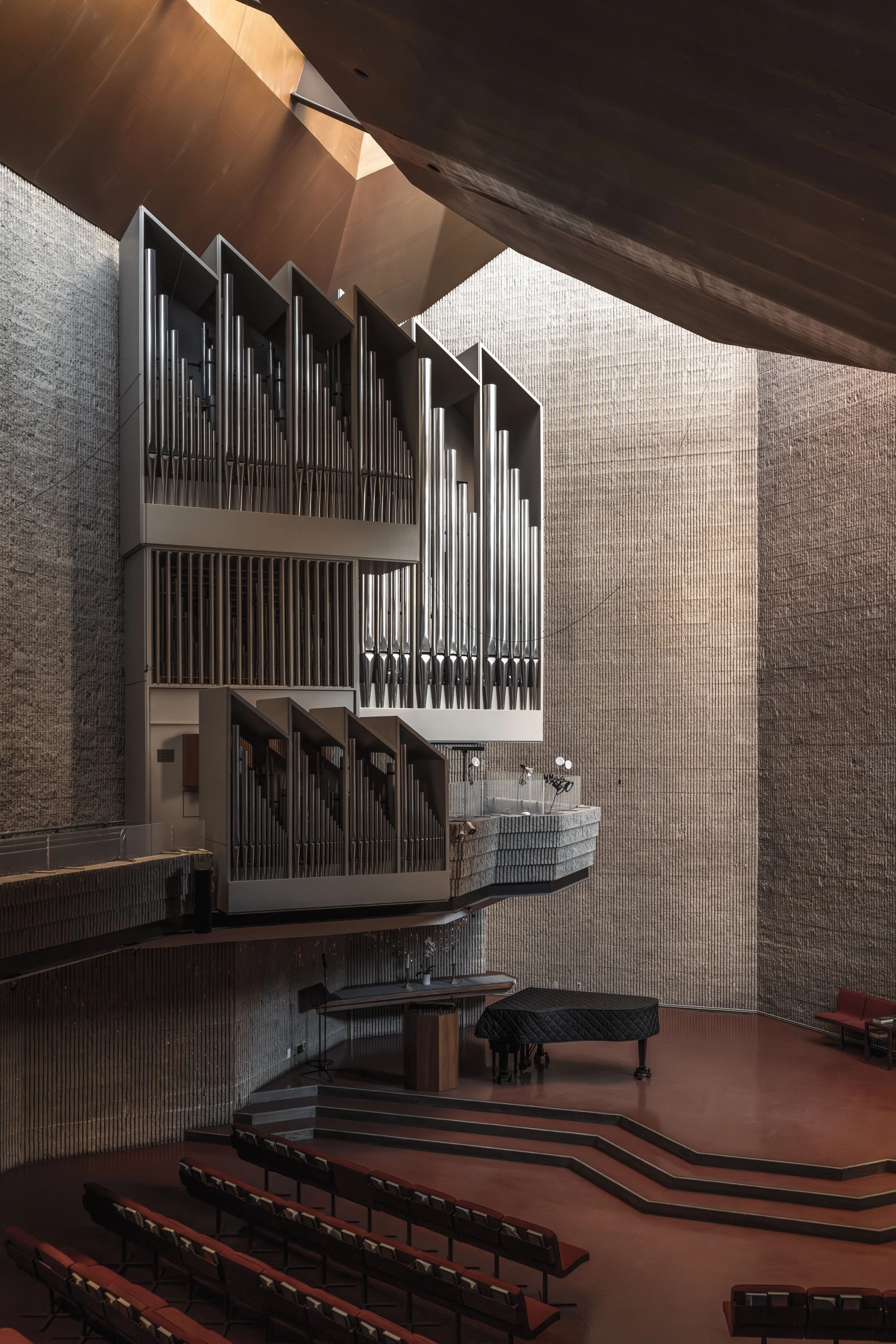 Interior view of a church sanctuary with a large pipe organ on an elevated platform, red cushioned choir seats, a grand piano, and stepped flooring, with textured wall surfaces and wood ceiling.