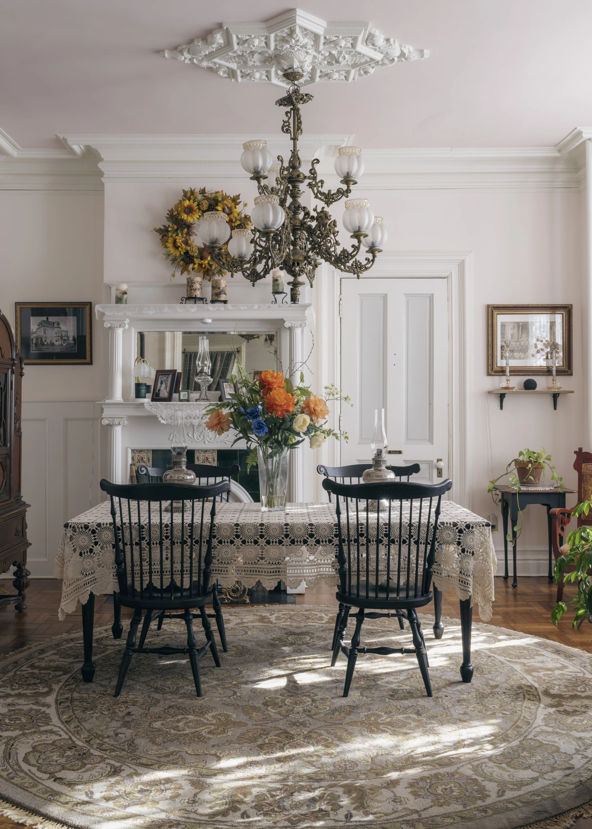 A dining room with a vintage chandelier, floral arrangements on a lace-covered table, a large mirror above the fireplace, and classic furniture and decor.