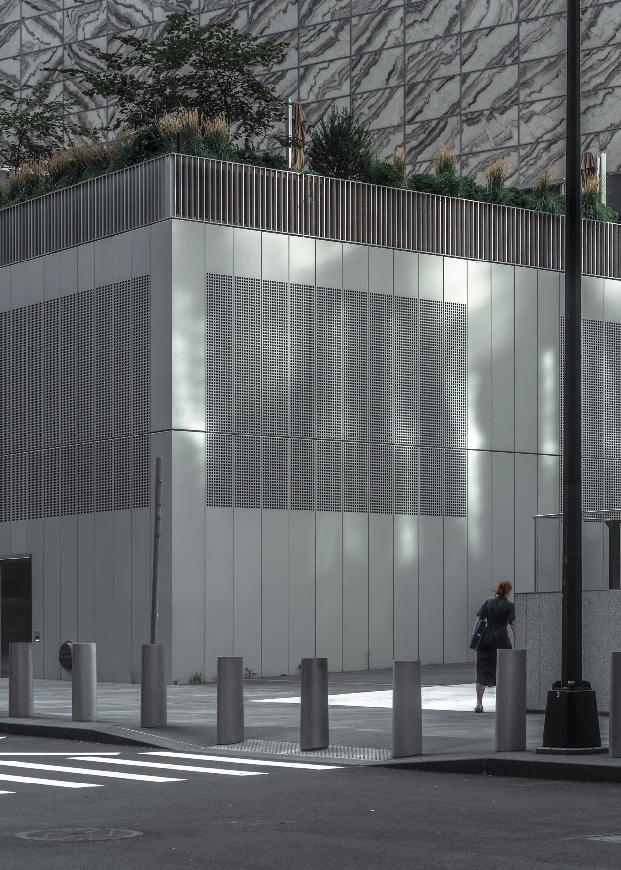 A woman walking past a modern building with metallic exterior panels and a landscaped rooftop garden with trees and plants.