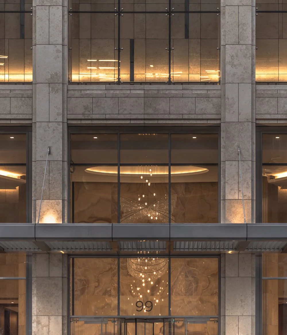 The facade of a modern building with large glass windows and stone walls, featuring a chandelier visible through the ground-floor window.