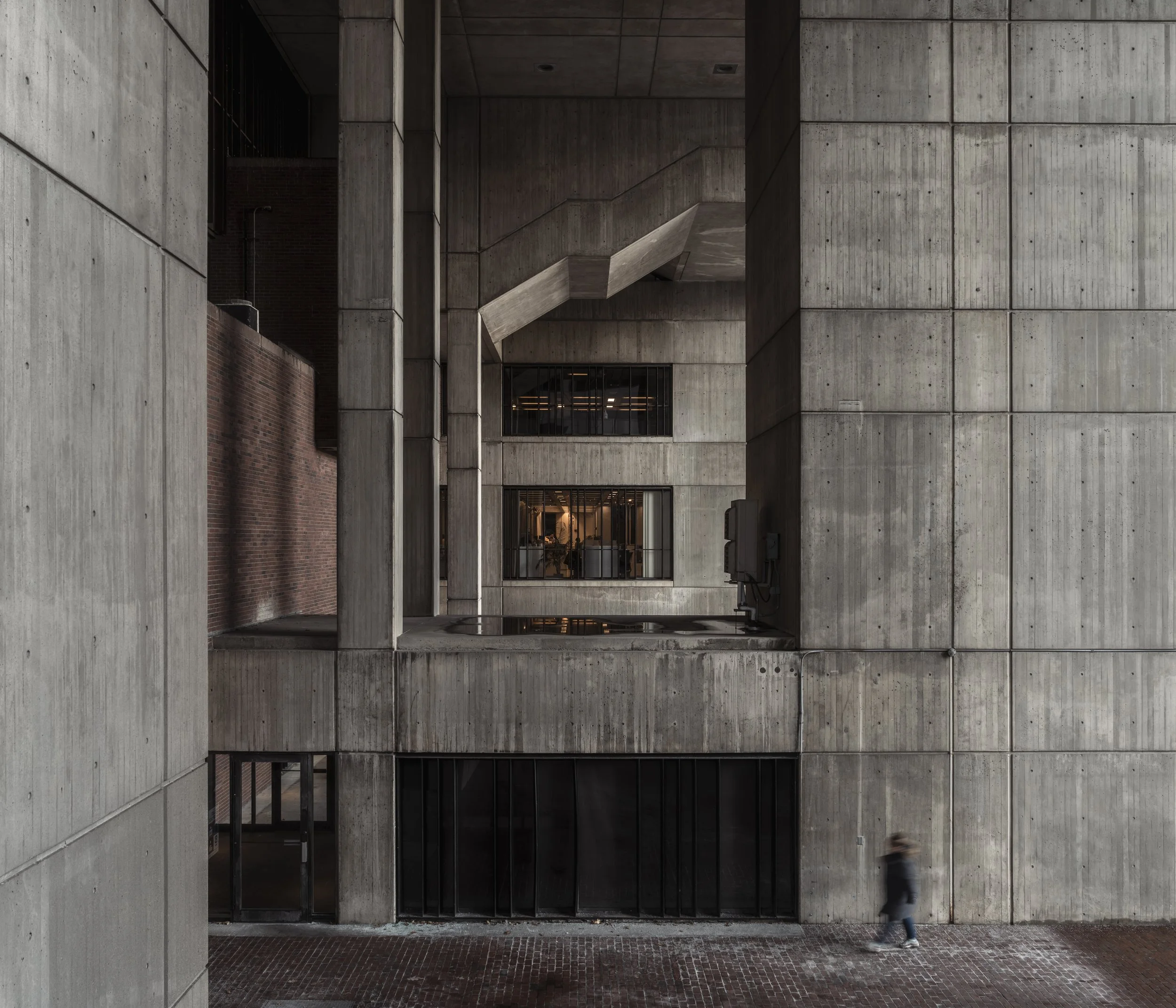 Modern concrete building with large rectangular windows, person walking on sidewalk in front, blurred motion.
