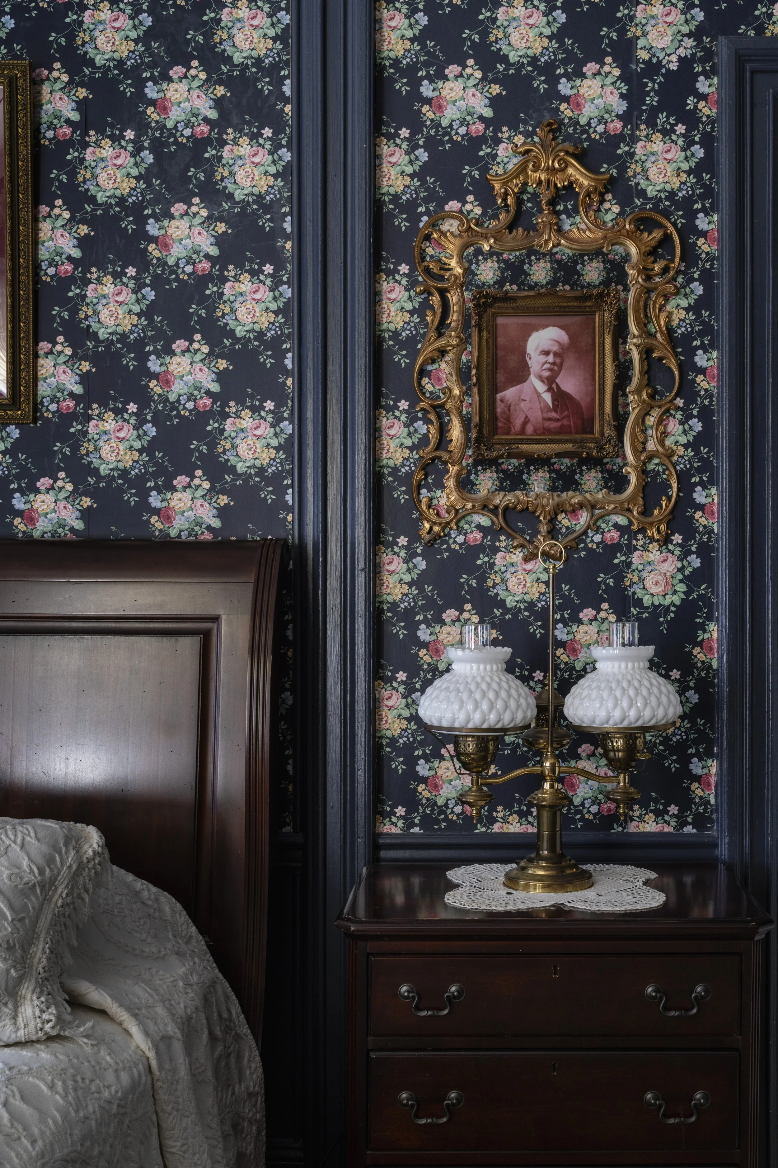 Vintage bedroom with floral wallpaper, wooden headboard, framed photo of an elderly person on the wall, ornate gold frame, and brass lamp with glass lampshades on a nightstand.