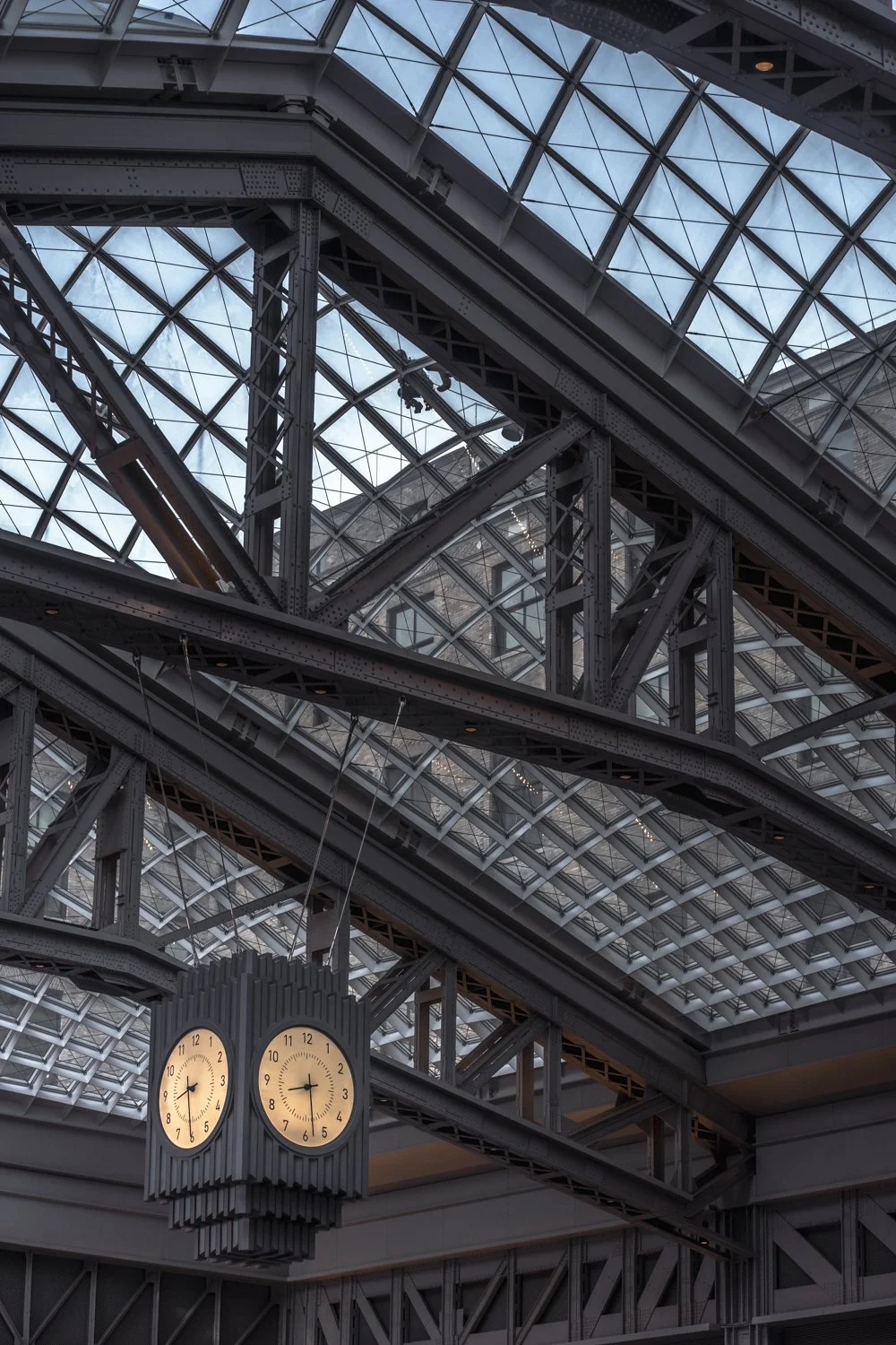 Interior view of a train station with steel beams and a glass ceiling, featuring a hanging clock showing 5:44.