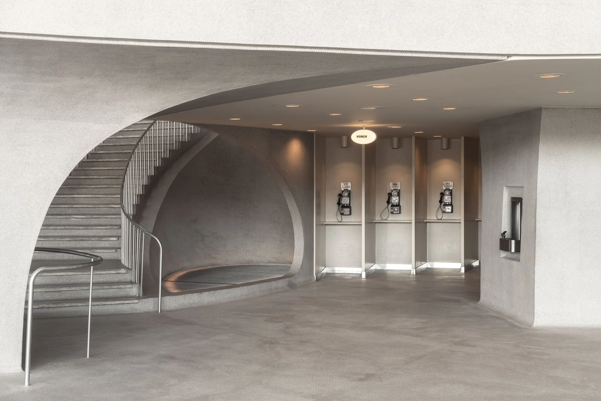 Public restroom area with a staircase on the left side, multiple payphones on the back wall, a women's sign overhead, and a vending machine on the right wall.