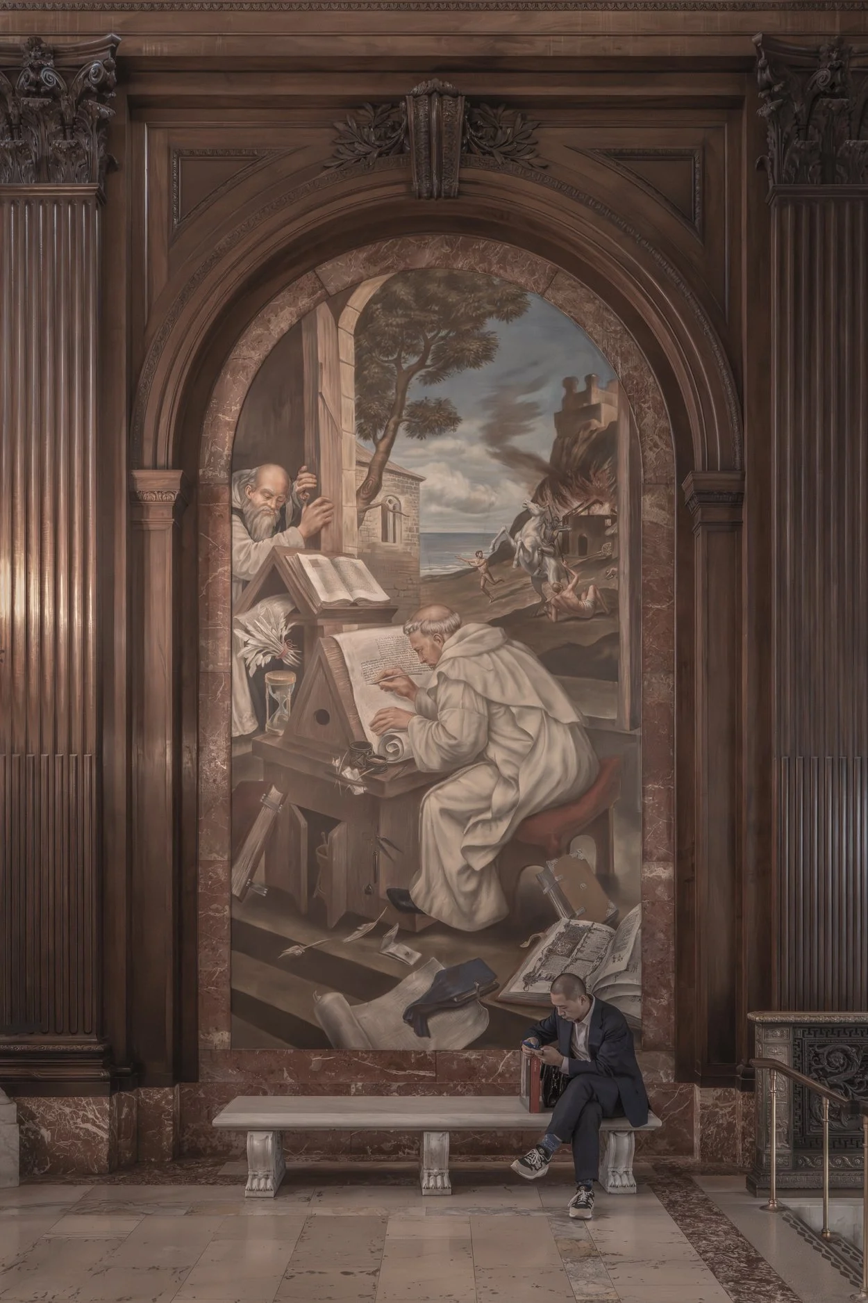 A large marble mural depicting a historical scene with a monk working at a desk, surrounded by classical architecture and mythological figures, set within an ornate wooden frame in a grand hall, with a man sitting on a bench looking at his phone belo