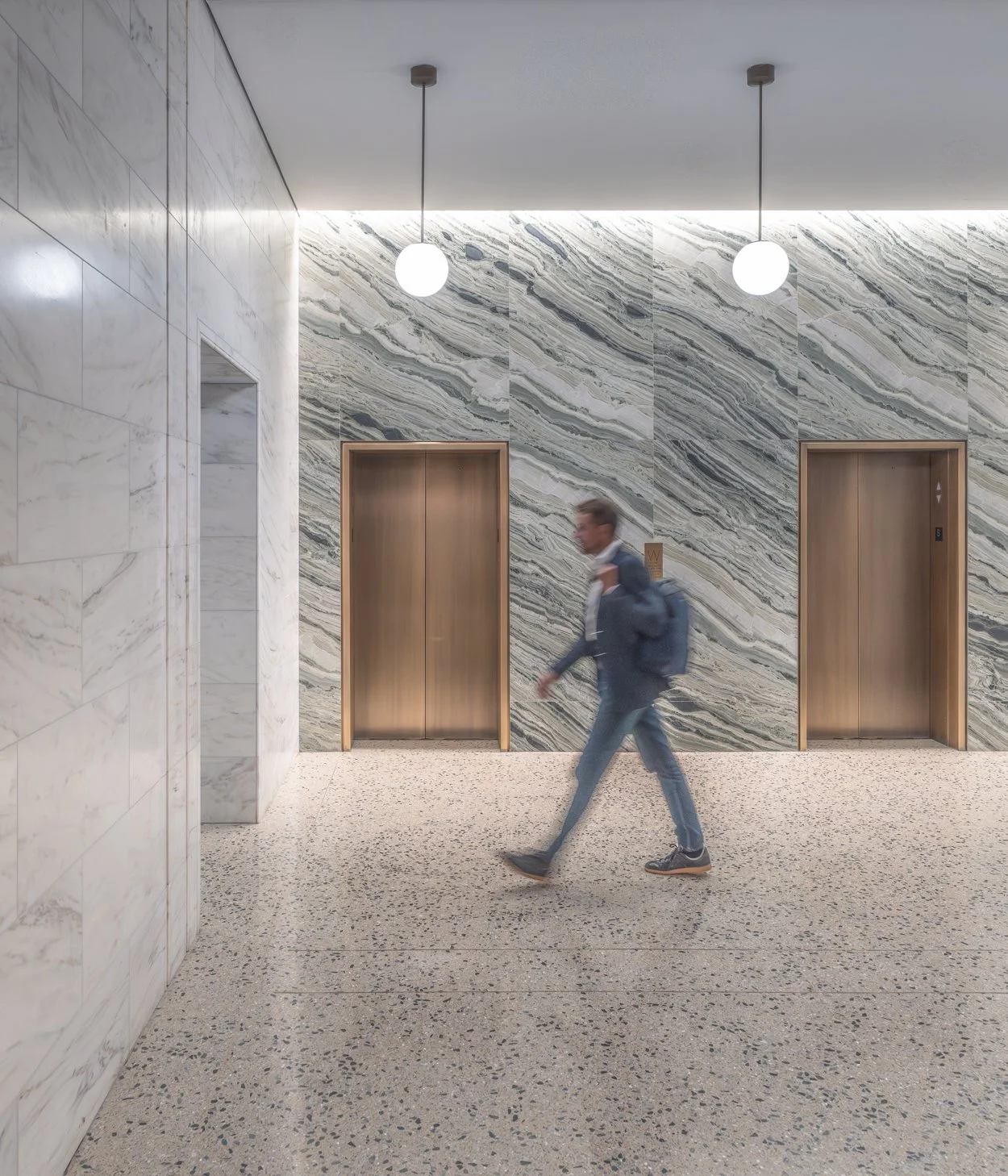 A person walking in front of two elevator doors in a modern building hallway with marble walls and a speckled floor, illuminated by round ceiling lights.