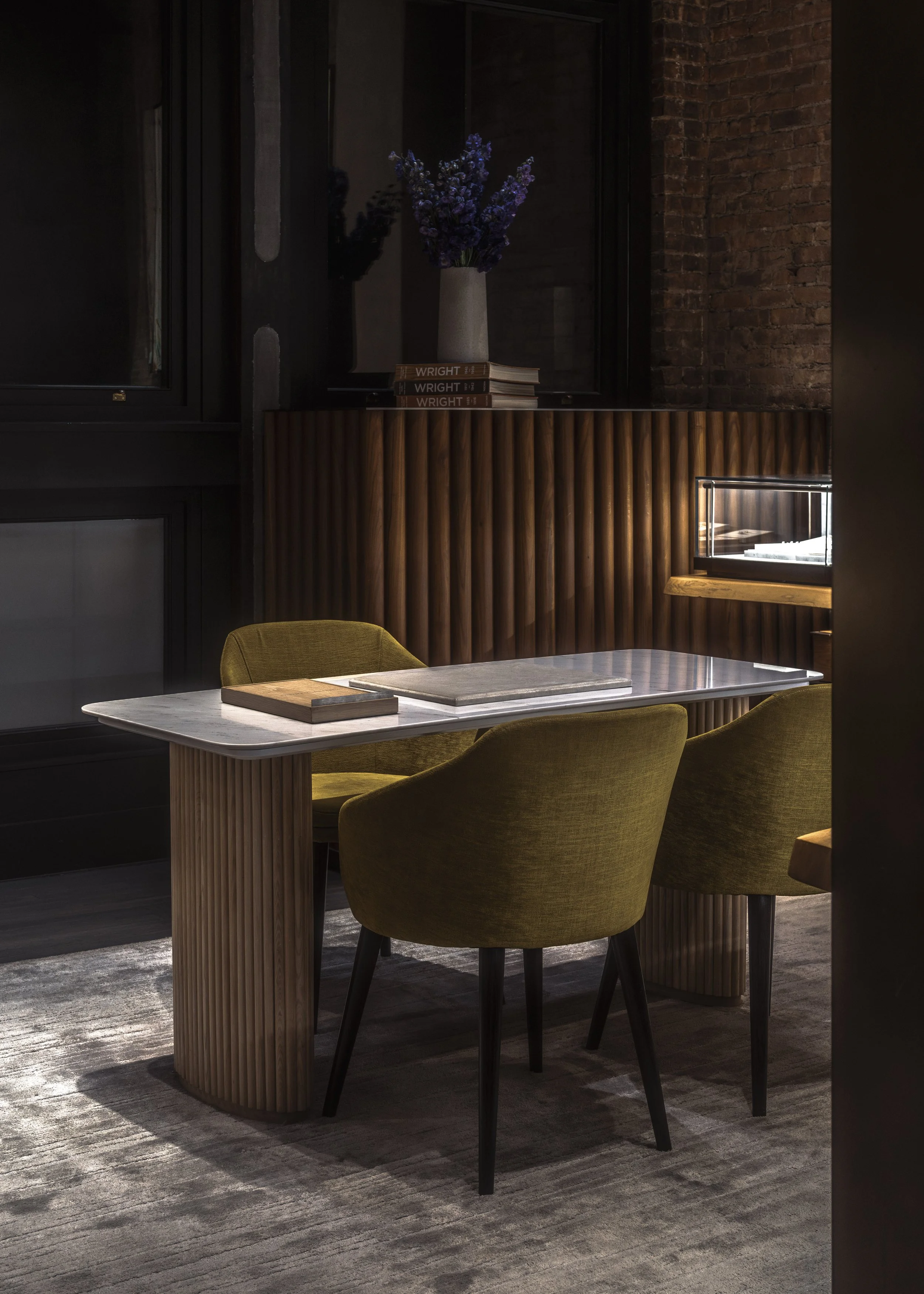 A modern dining area with a marble-topped table, two olive-green upholstered chairs, a wooden sideboard with vertical slats, a small glass display case, a stack of books and a tall vase with purple flowers on top.