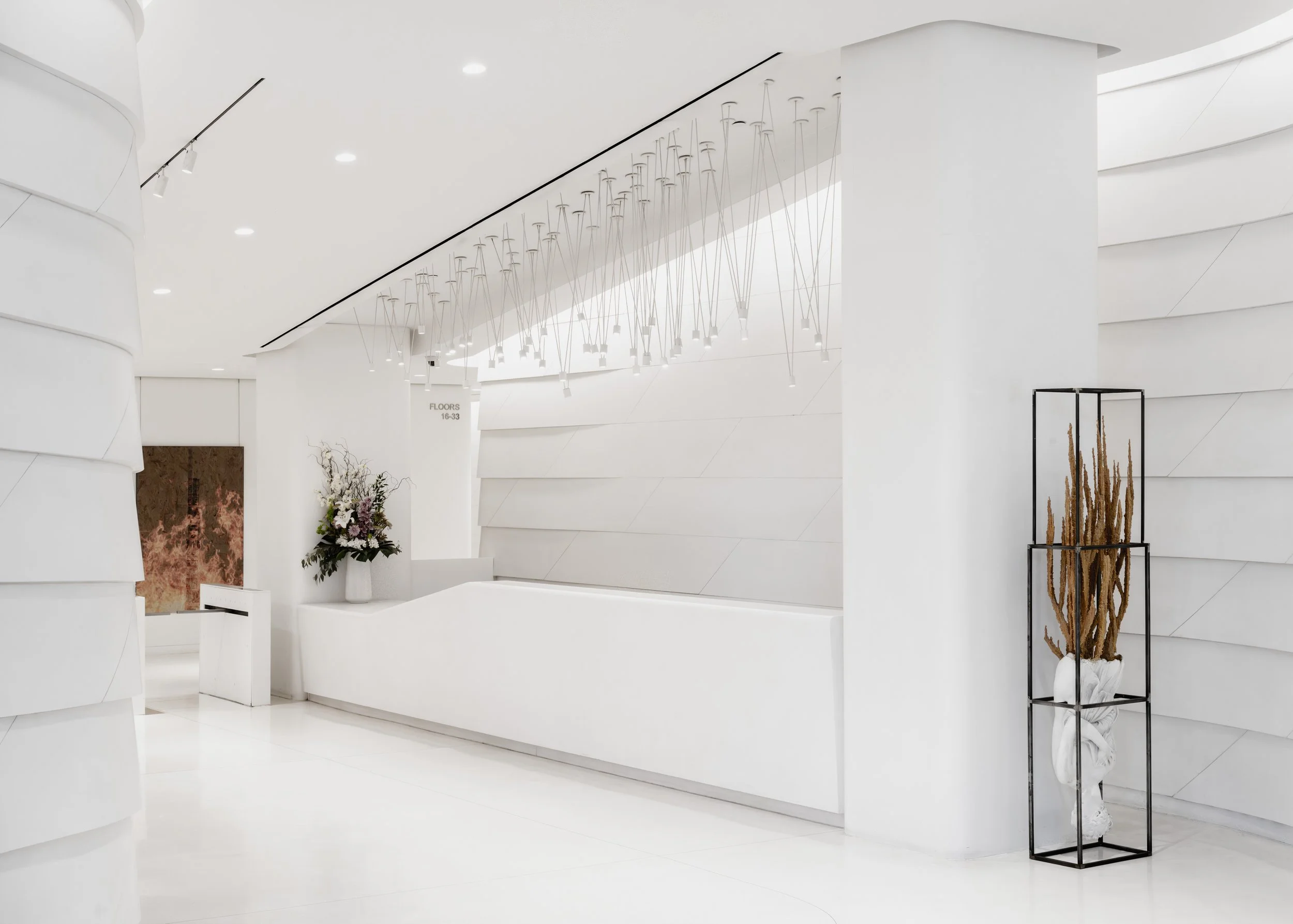 Minimalist modern white lobby with a curved wall, tall white reception desk, flower arrangement, modern chandelier, and a black metal shelf holding a sculpture with dried plants.