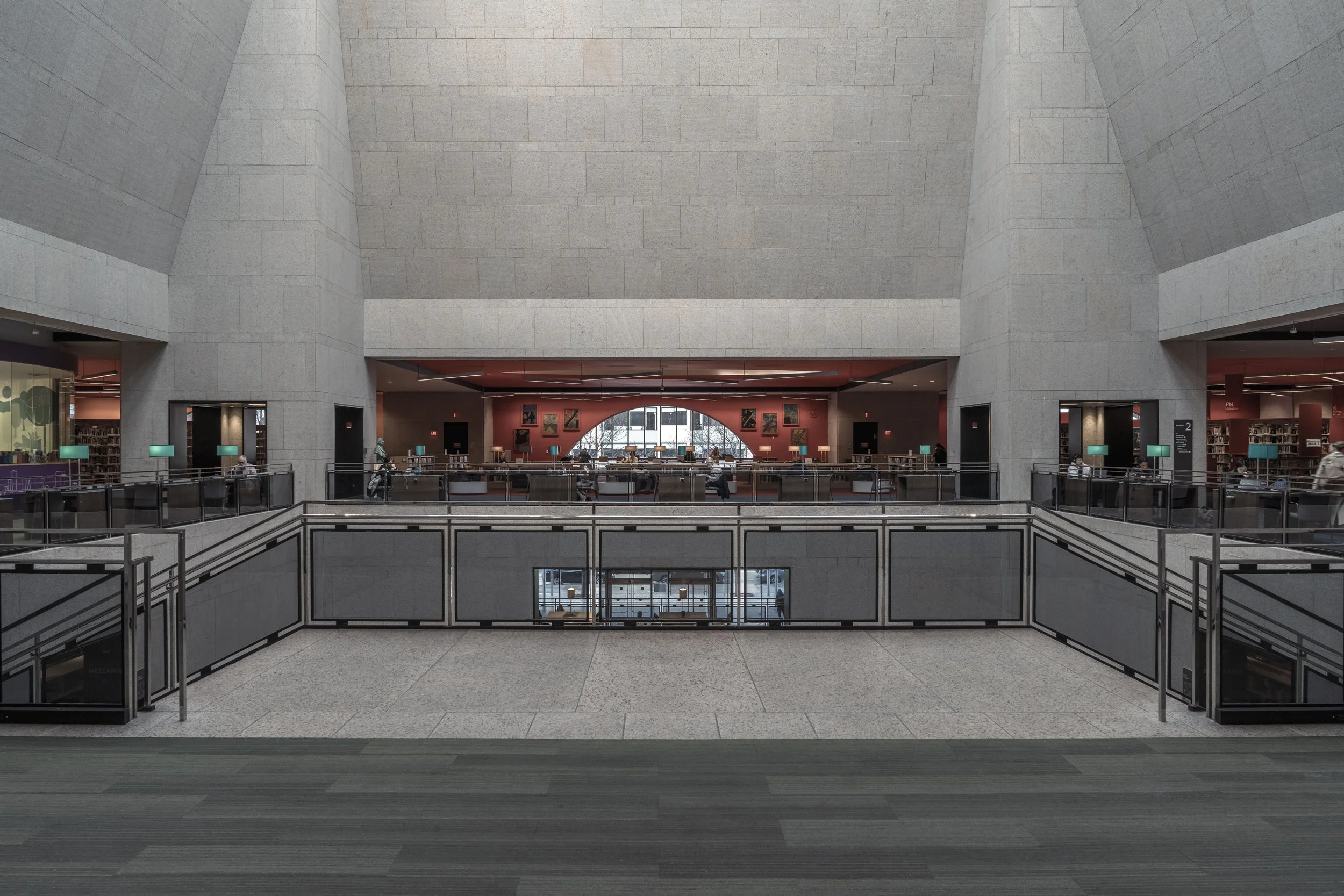 Interior of a modern library with a large open space, multiple levels, and a red-colored wall behind a central seating area with tables and chairs. Walls are made of stone, and there are several teal table lamps and artwork on the walls.