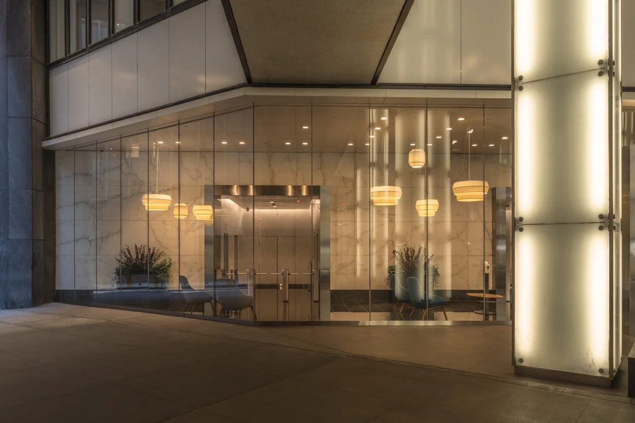 Nighttime view of a modern building entrance with glass walls, marble walls inside, hanging pendant lights, and contemporary chairs and plants visible through the windows.