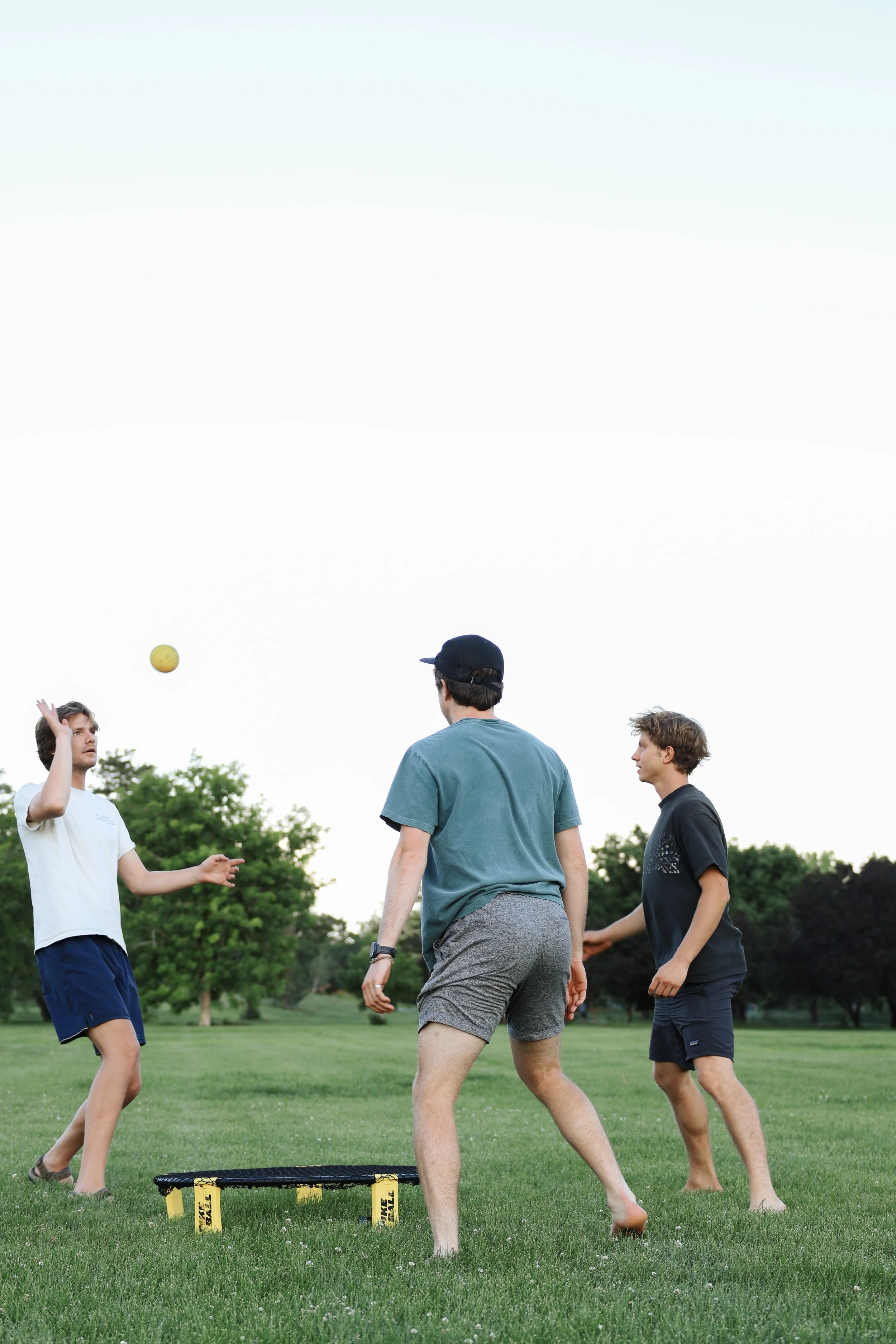 Three young men playing Spikeball on a grassy field outdoors, with one preparing to hit a yellow ball, a small black and yellow net on the ground, and trees in the background