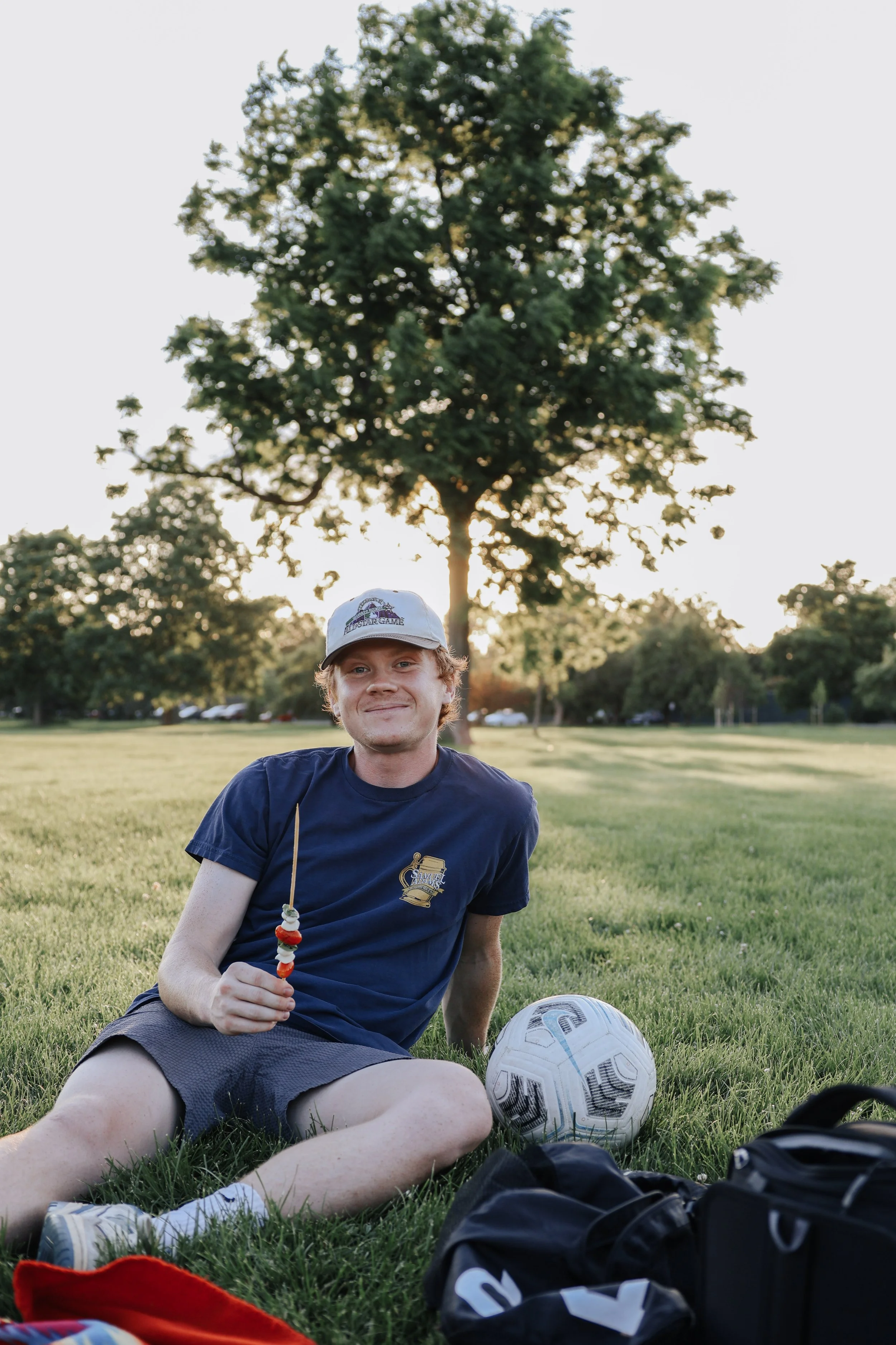 A young man sitting on grass in a park holding a skewer of food with a soccer ball beside him, with trees and parked cars in the background.