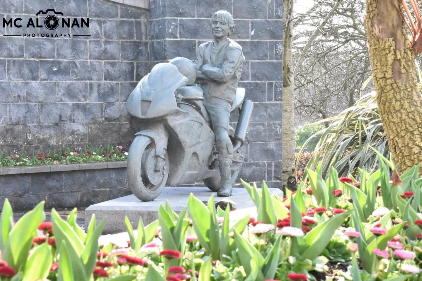 Statue of a person leaning on a motorcycle, surrounded by flowers