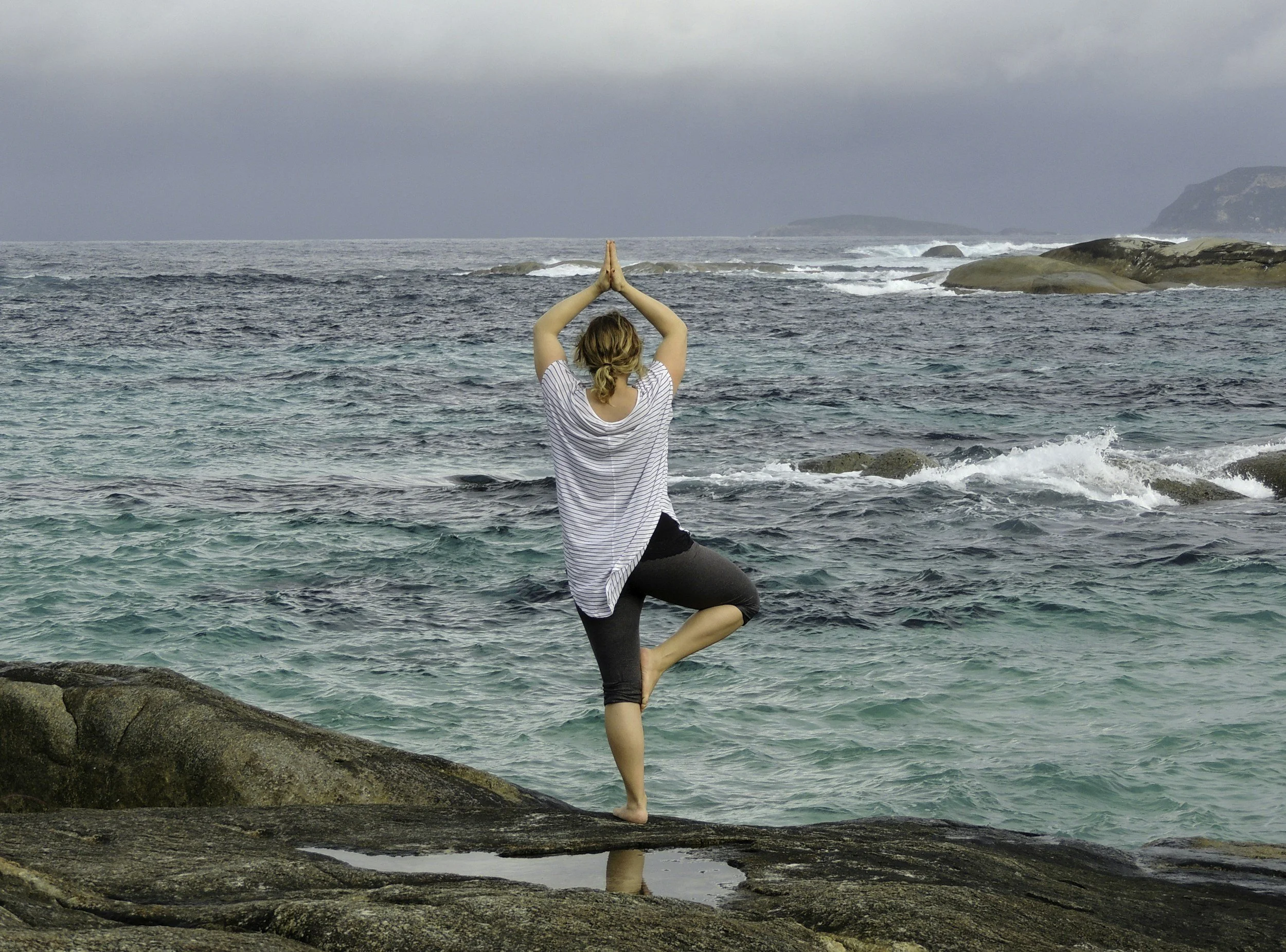 Person practicing yoga in tree pose on rocky coast overlooking the ocean under a cloudy sky. Pose represents balance training.