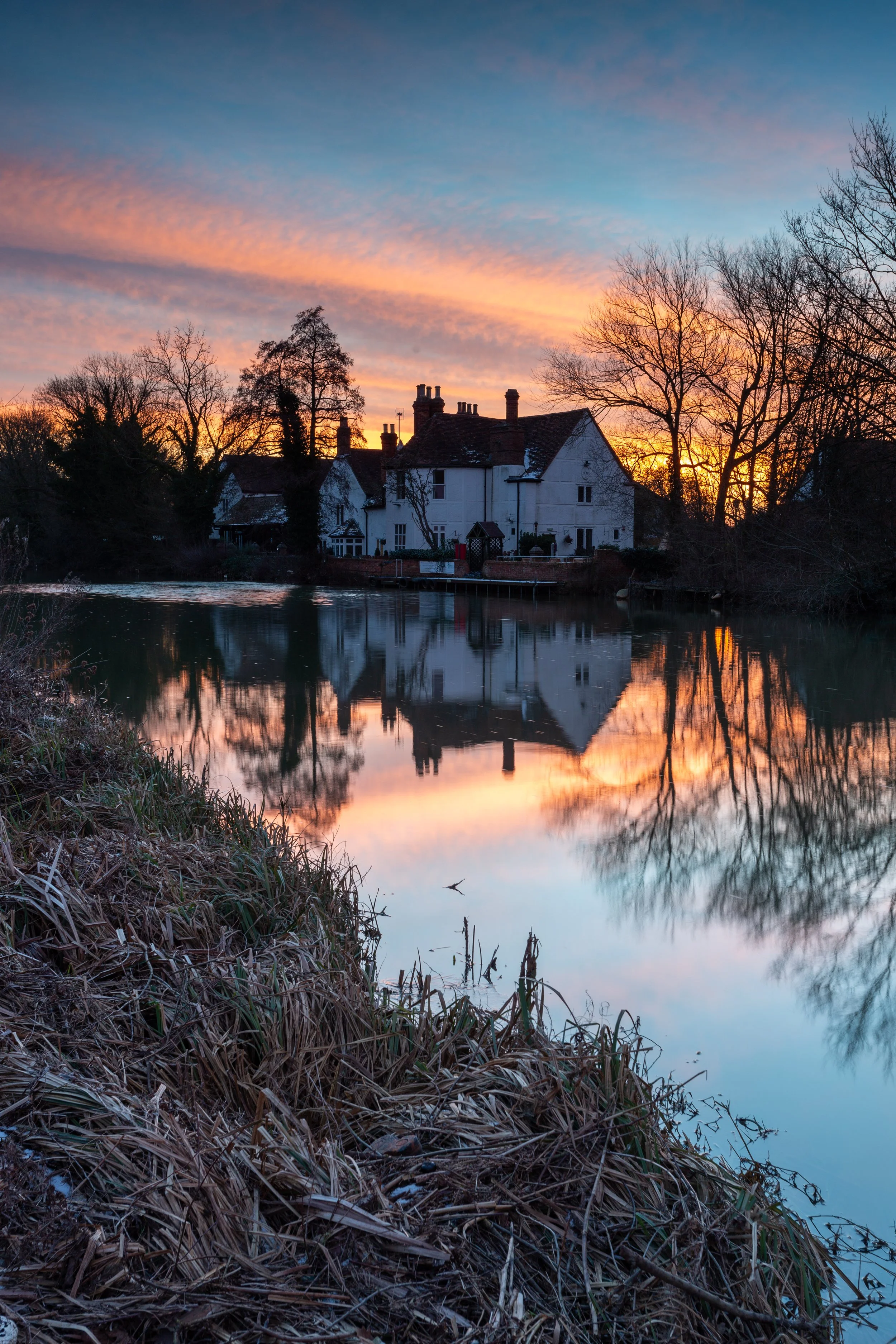 A sunset over a house reflecting in a calm river, surrounded by leafless trees and dry grass in the foreground.