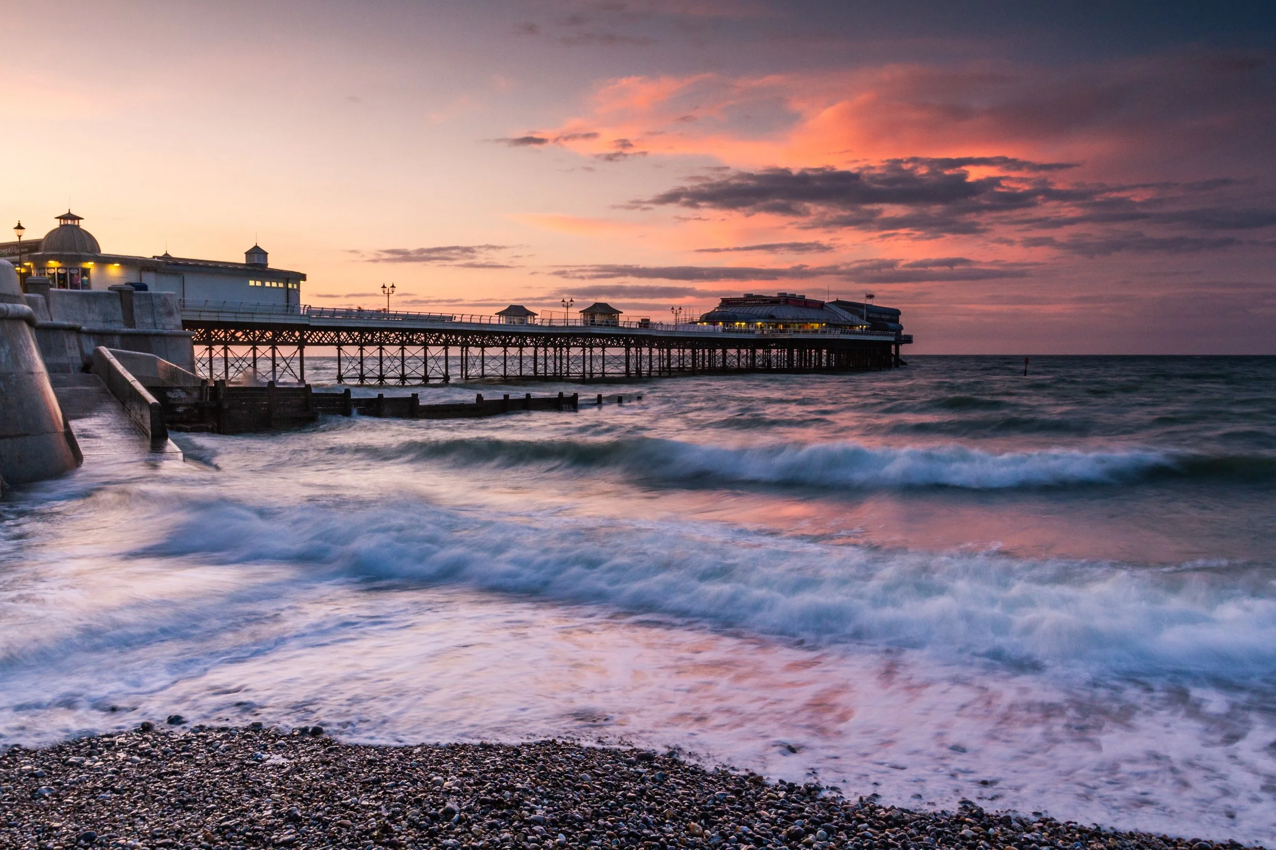 Sunset view of a pier extending into the ocean with waves crashing on a pebble beach in the foreground.