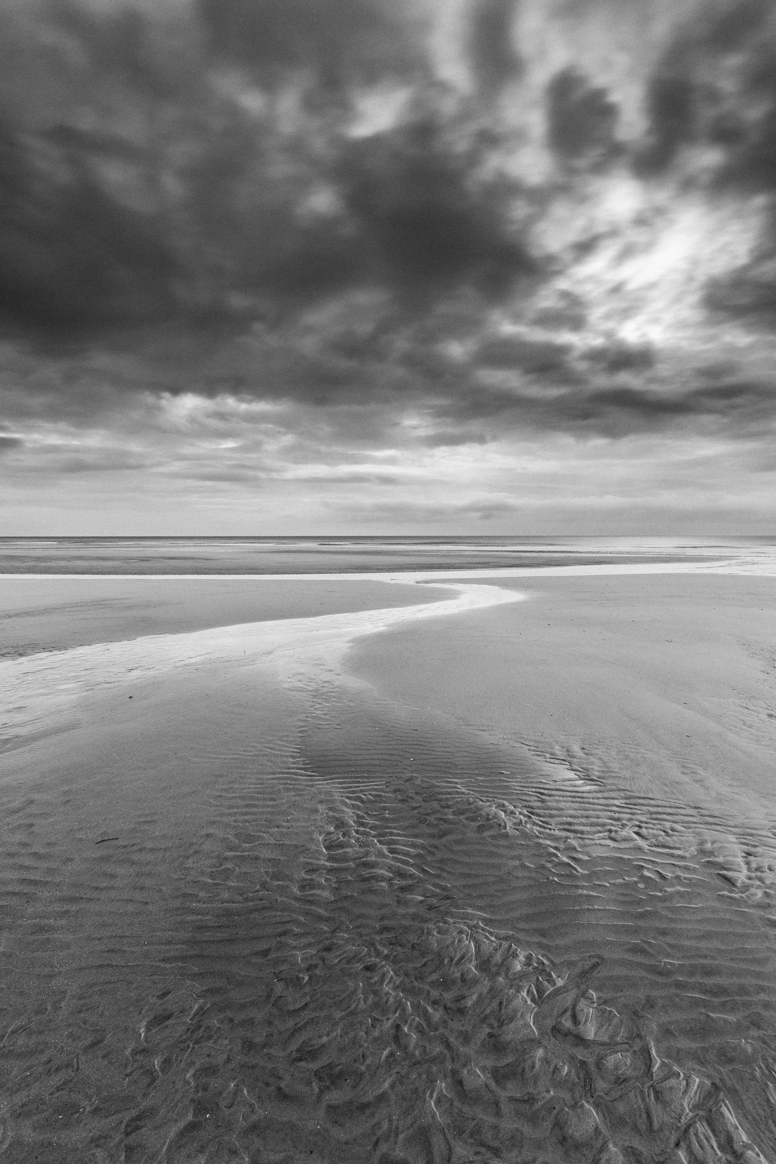 Black and white photograph of a deserted beach with a winding stream of water, cloudy sky, and calm ocean in the distance.