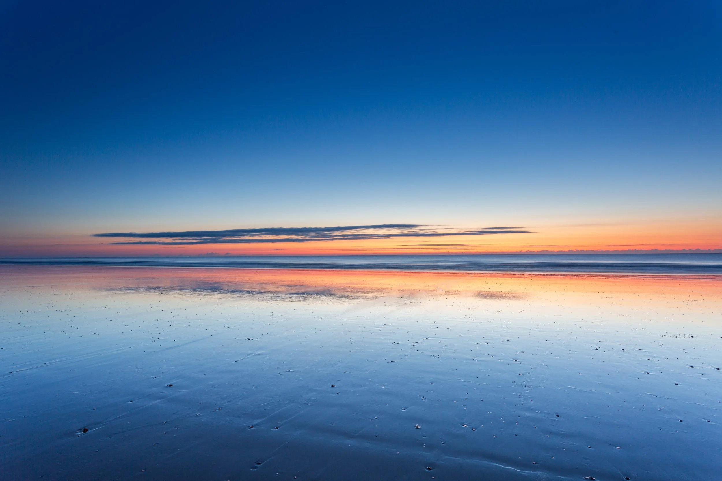 Serene beach at sunset with a colorful sky, calm water reflecting the sky, and footprints in the wet sand.