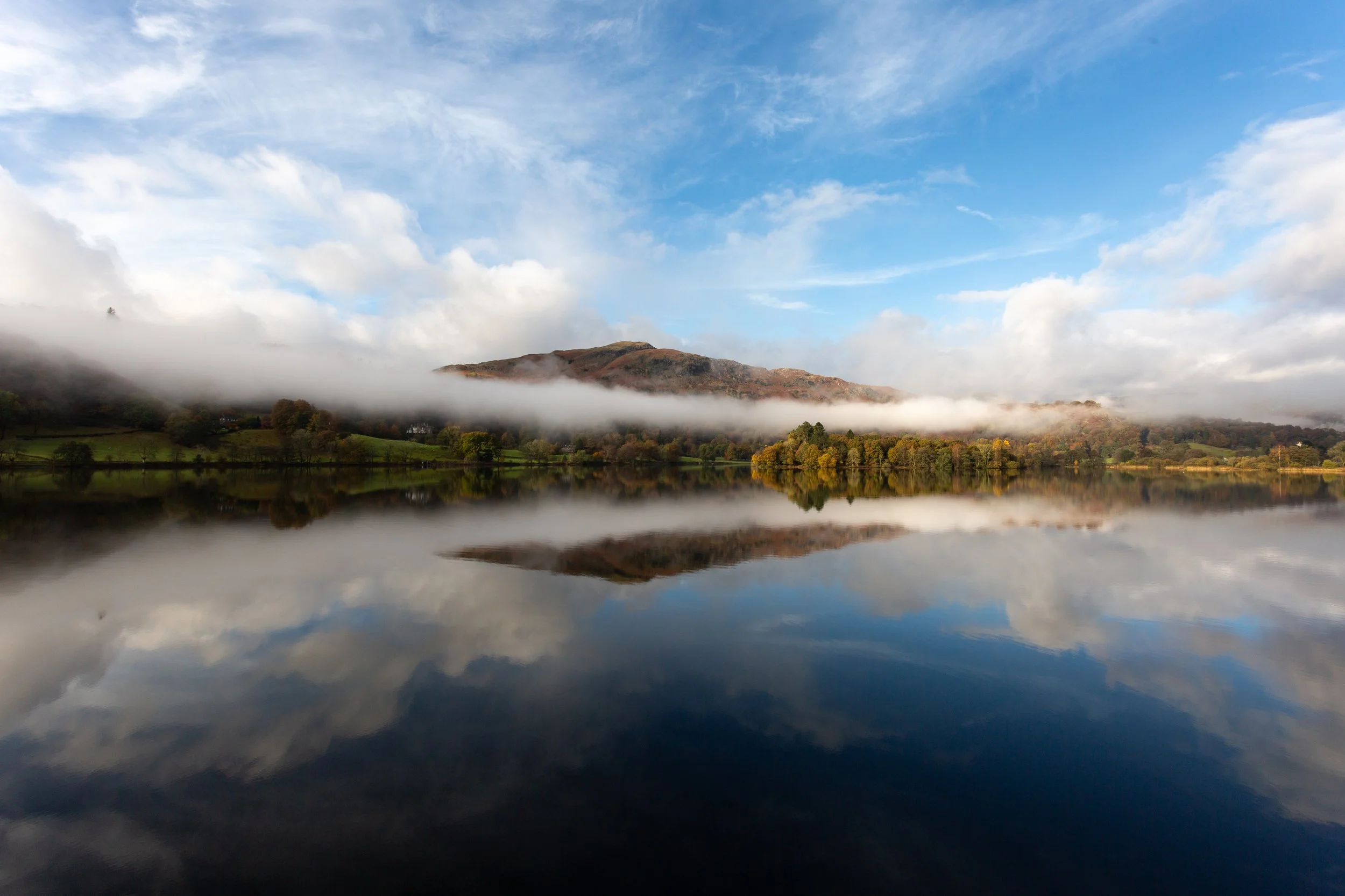 Landscape of a mountain range reflected in a lake with a partly cloudy sky and some fog over the mountains.