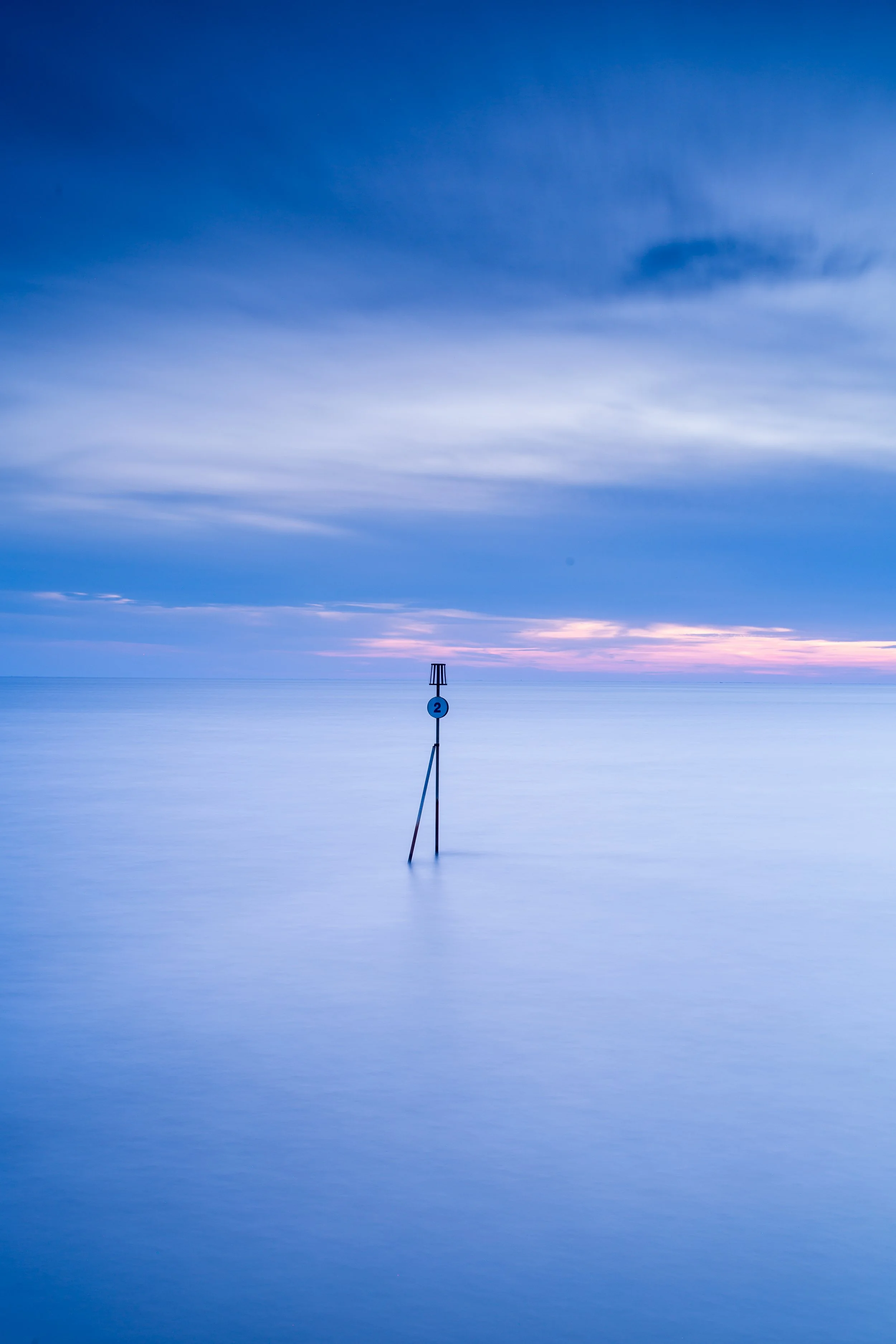 A calm sea at dusk with a sky in shades of blue and purple, featuring a navigation marker with a blue circle and the number 2, partially submerged in the water.
