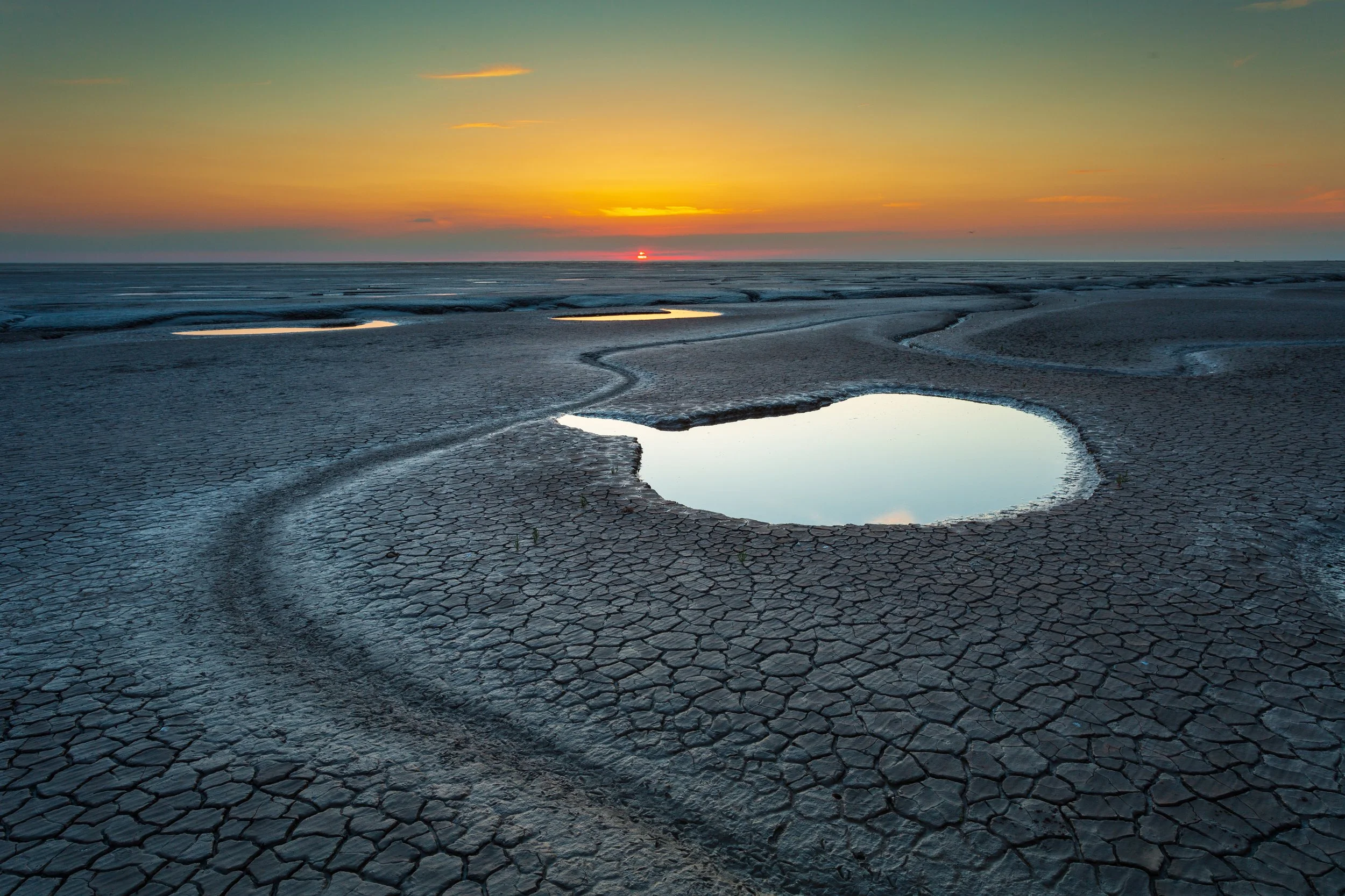 Sunset over a cracked, dry landscape with small water-filled craters reflecting the sky.