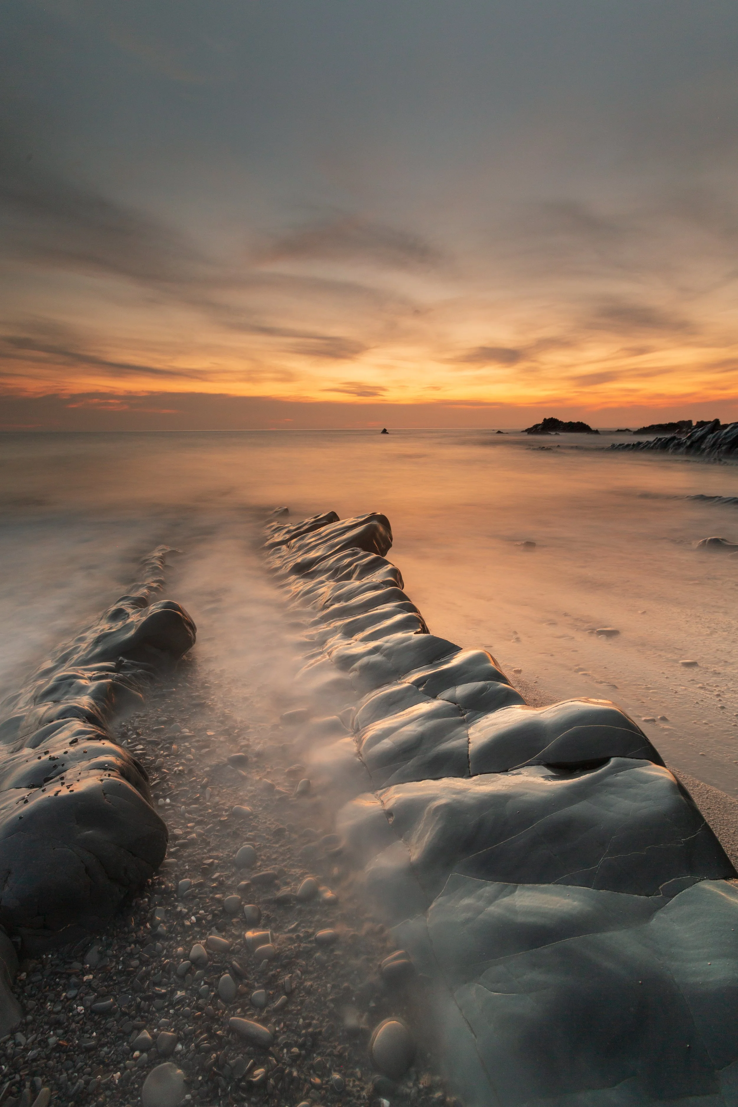 Sunset over the ocean with a rocky breakwater extending into the water, calm waves, and a partly cloudy sky.