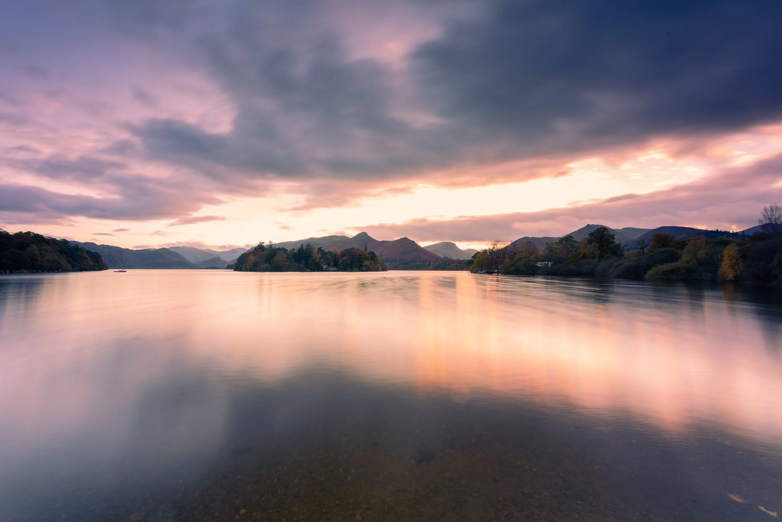 Calm lake surrounded by mountains and trees, under a colorful sunset sky with pink, purple, and orange clouds.