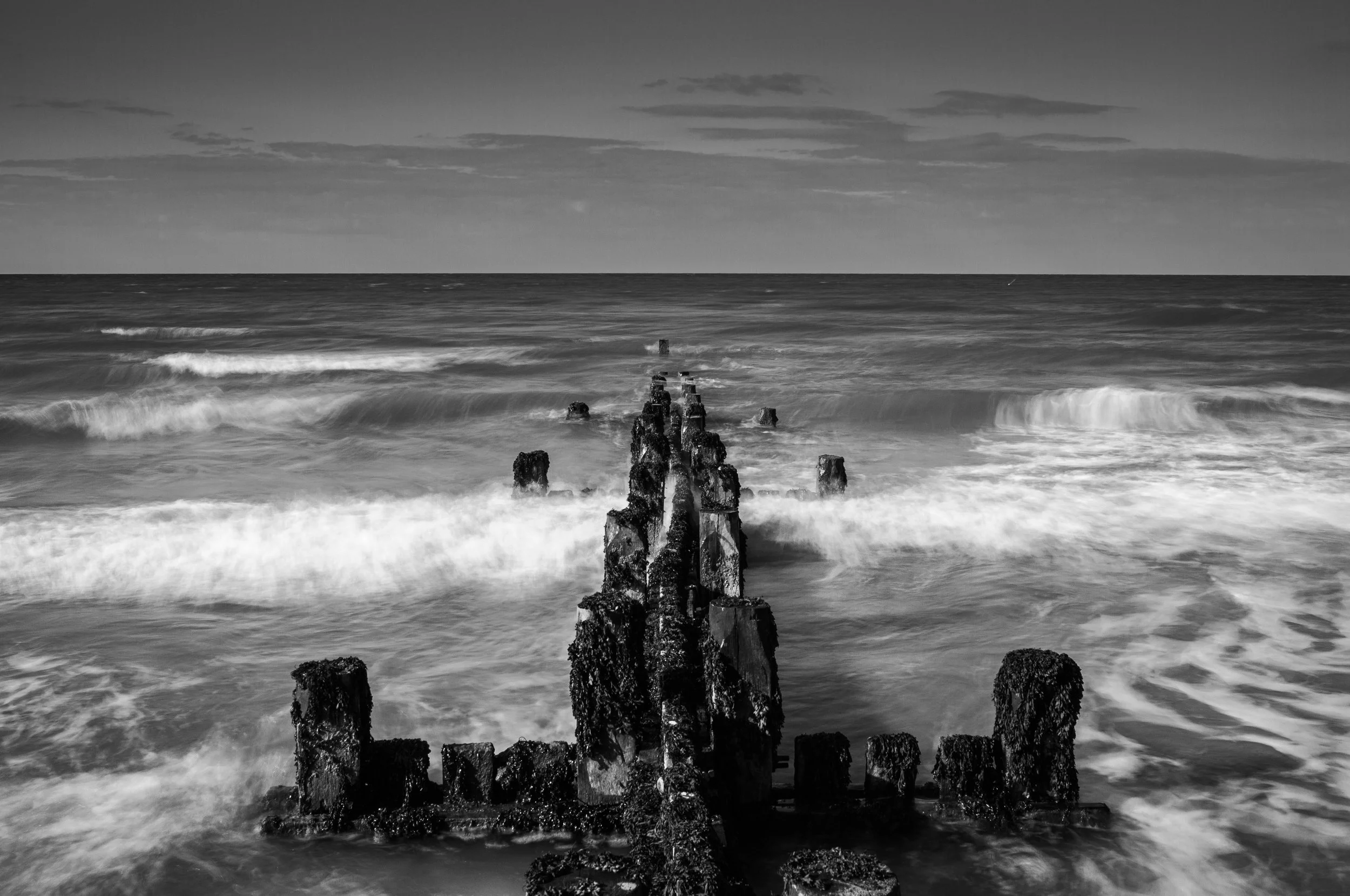 Black and white photo of water crashing against old, weathered wooden posts of a pier or breakwater in the ocean, with clouds in the sky above.