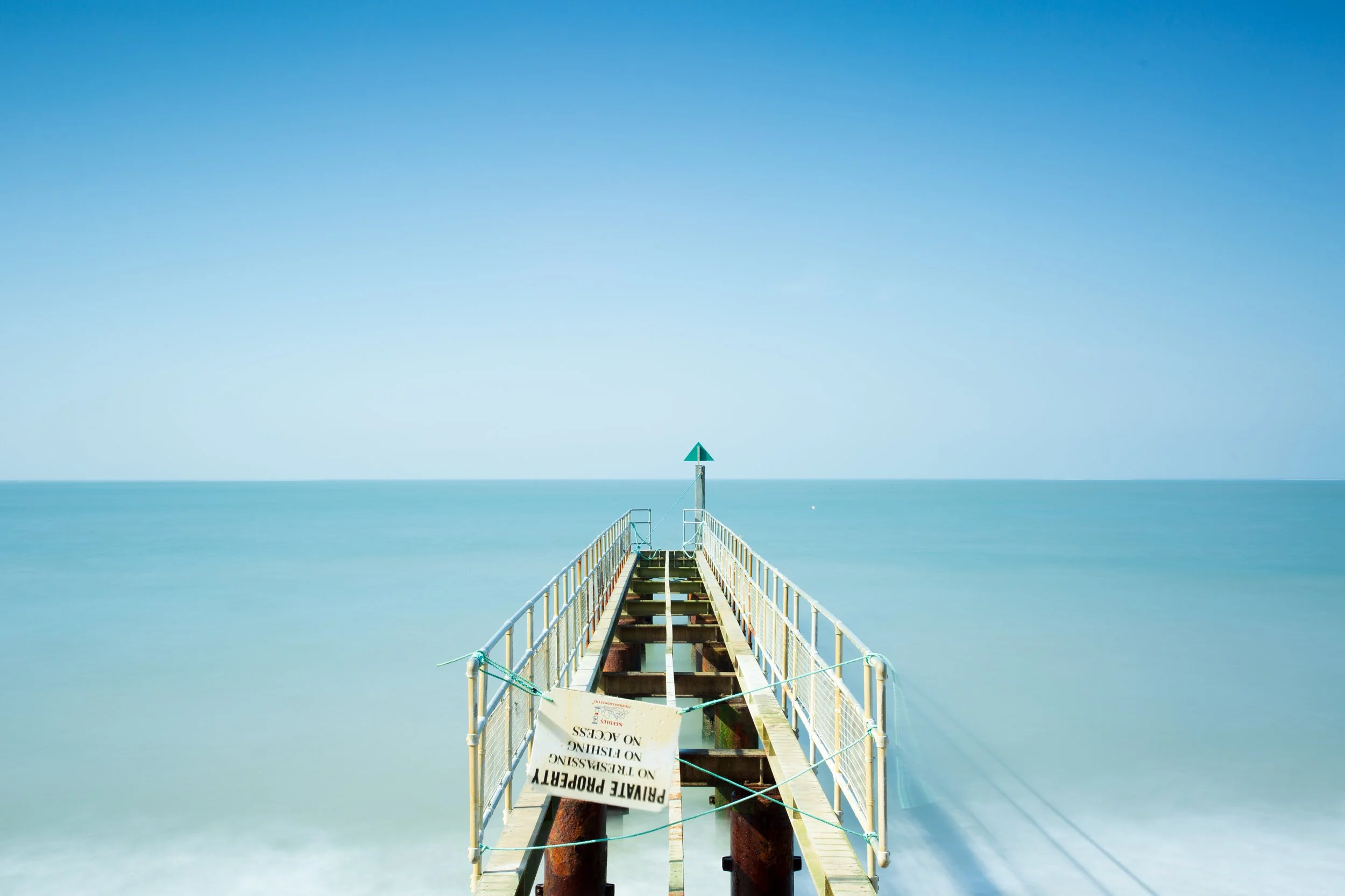 A wooden pier extending into a calm ocean with a blue sky, with a sign warning not to trespass, cautioning against climbing on the pier.