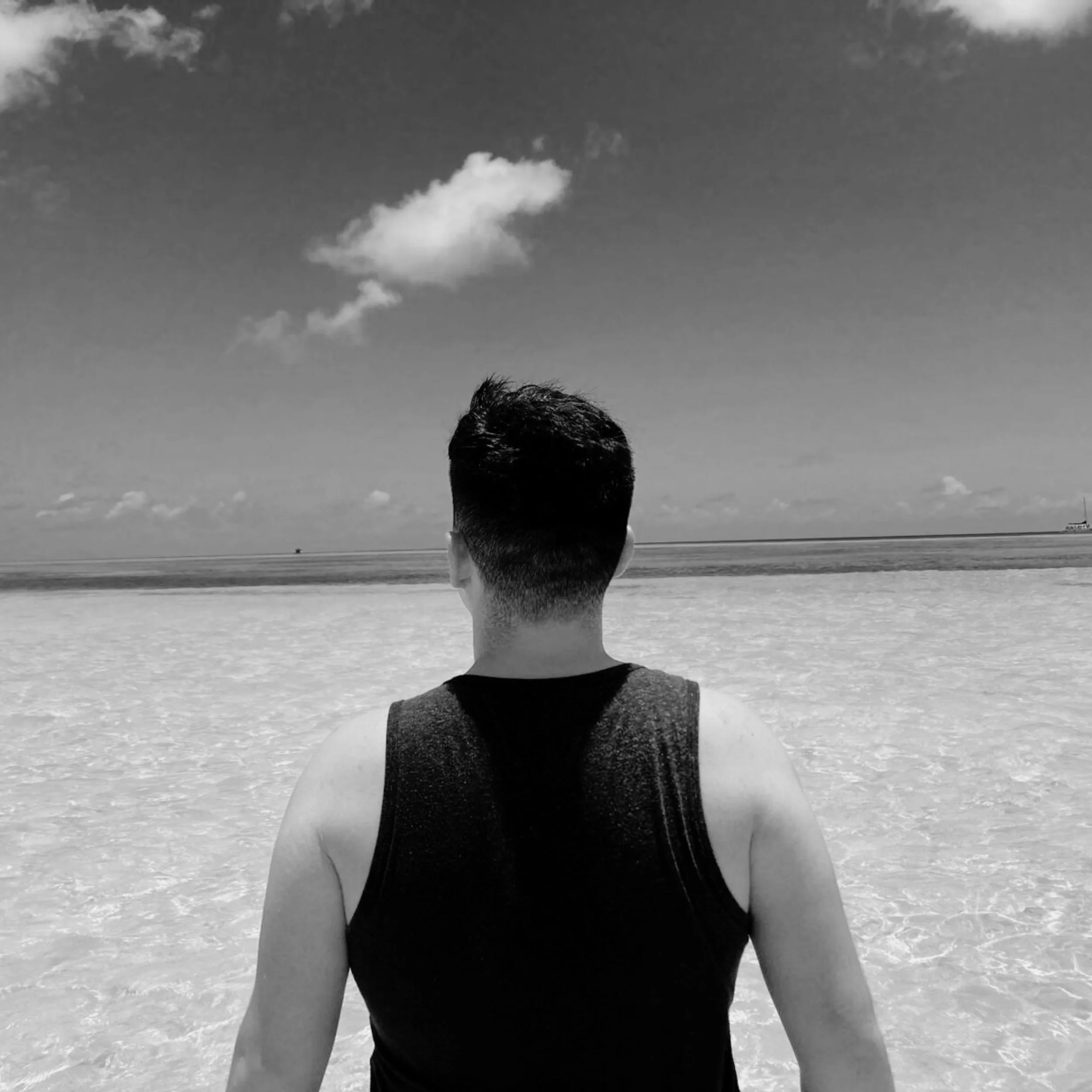 Hombre de espaldas con camiseta sin mangas en la playa mirando hacia el mar y un cielo nublado.