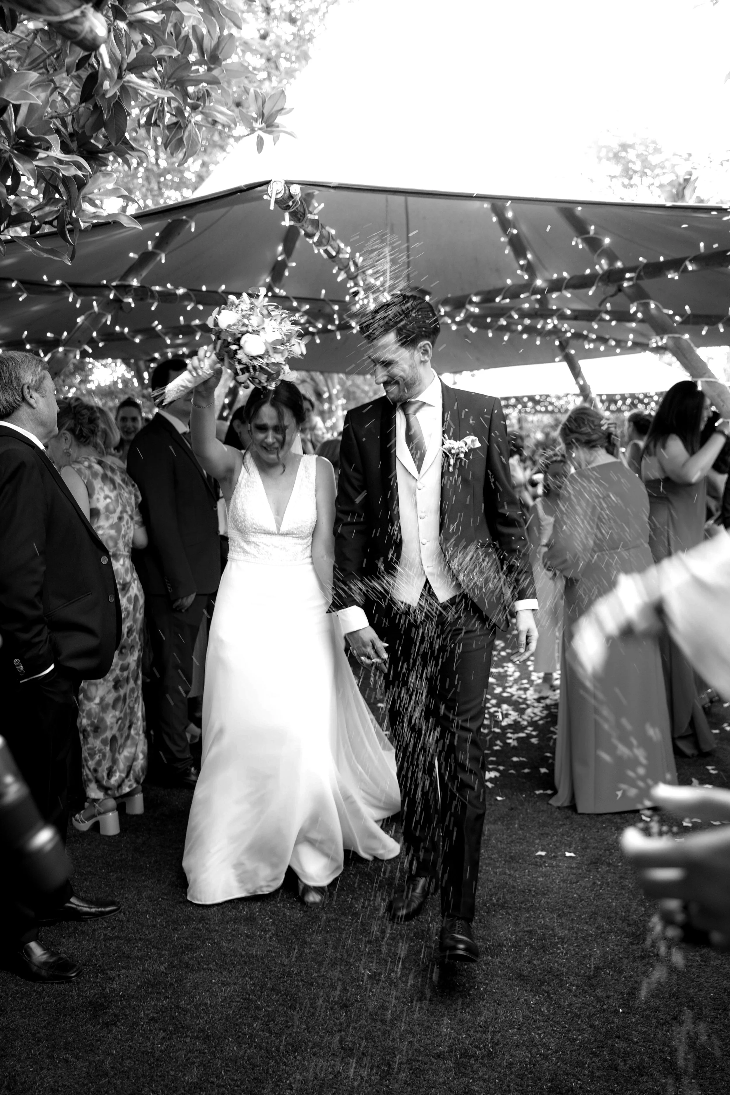 Una pareja de recién casados caminando entre invitados en una boda al aire libre, con ella lanzando flores y él sonriendo, mientras llueven confeti.