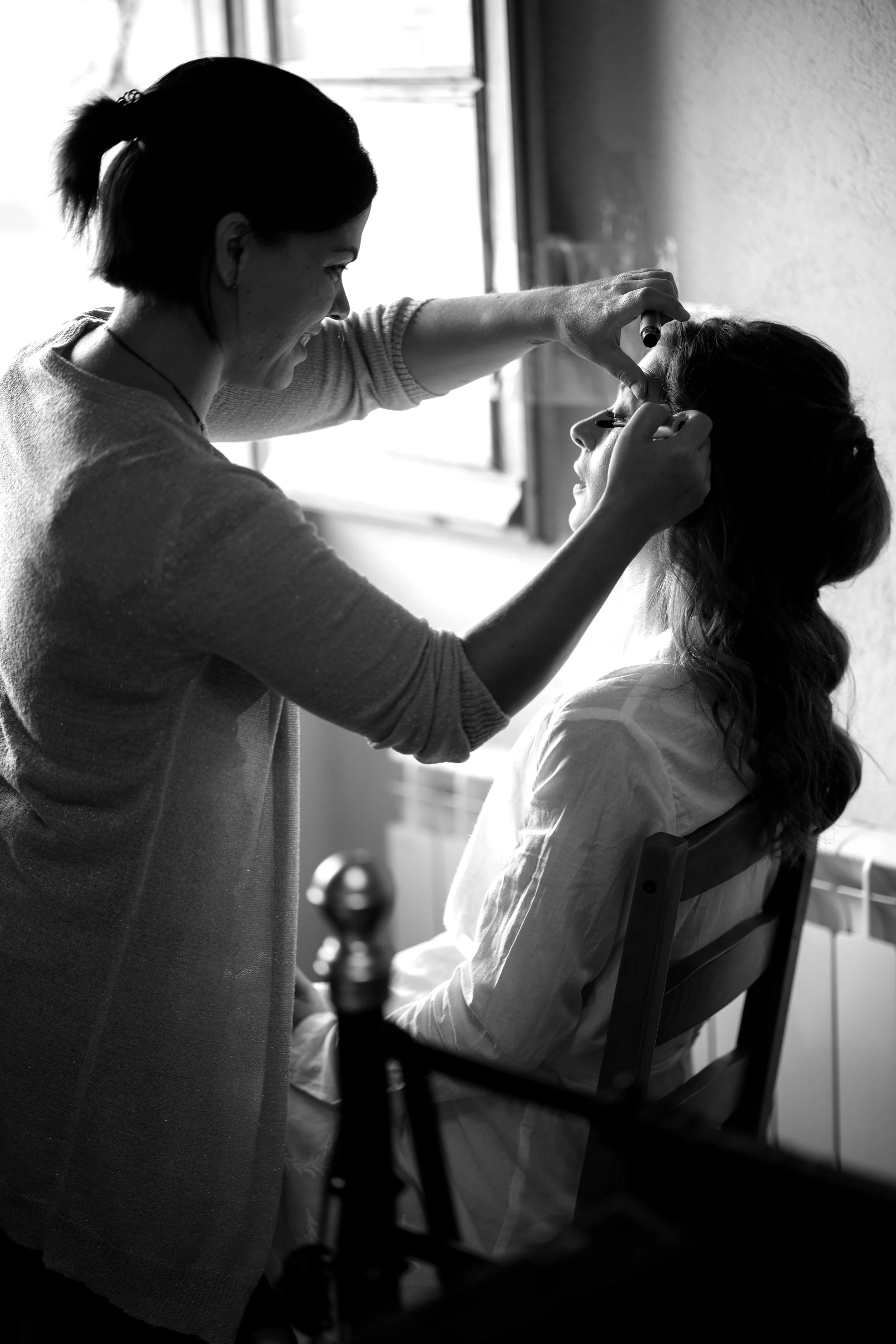 Una mujer maquillándose a otra mujer que está sentada en una silla junto a una ventana