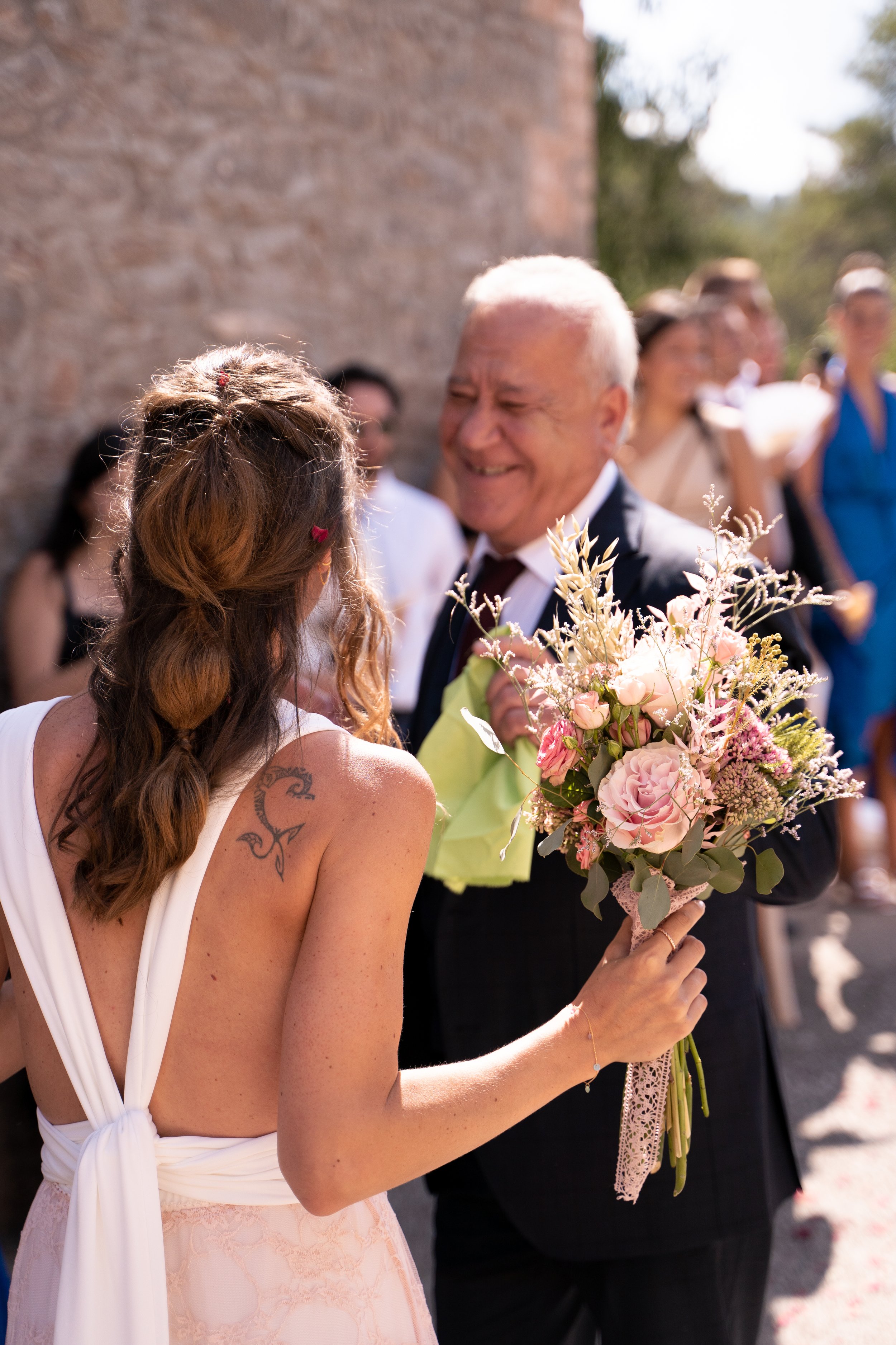Una mujer con vestido blanco y cabello rizado entregando un ramo de flores a un hombre mayor en un evento al aire libre.