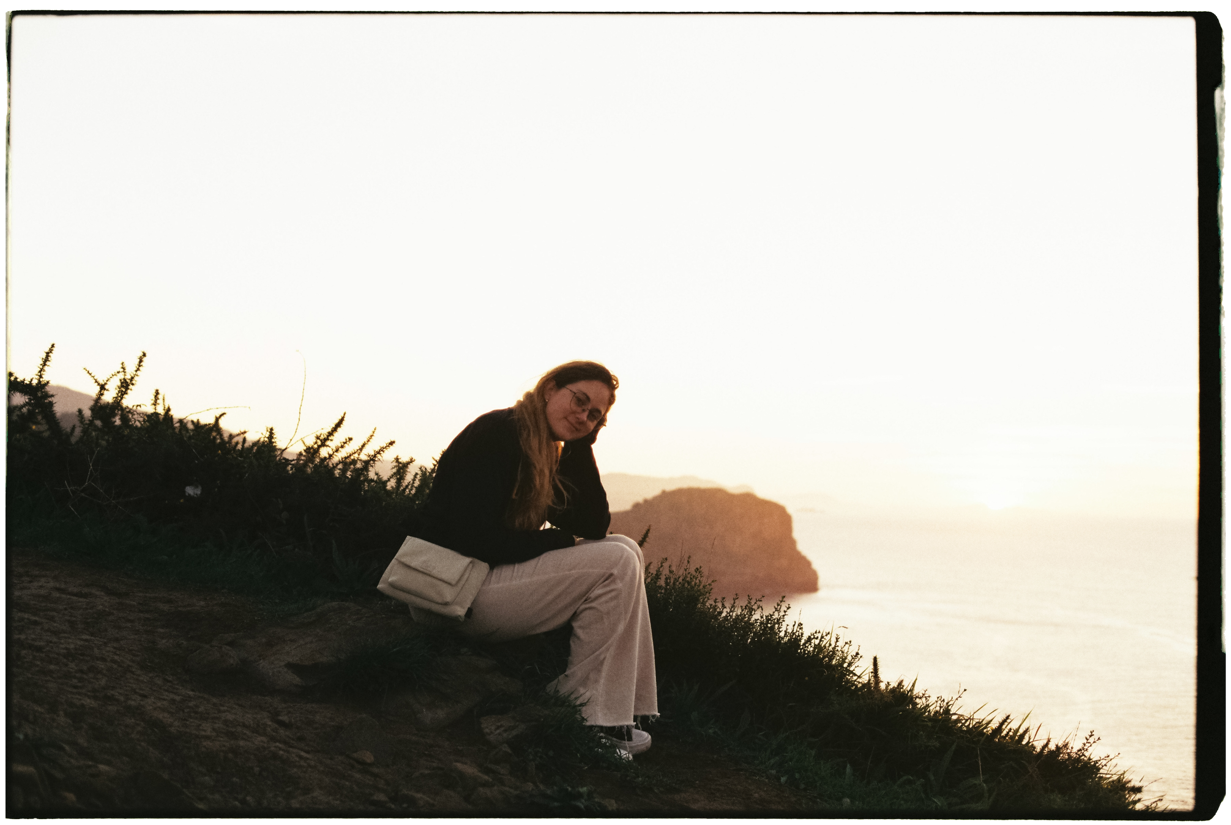 Mujer sentada en la orilla de un acantilado durante el atardecer con vista al mar y rocas en el fondo.