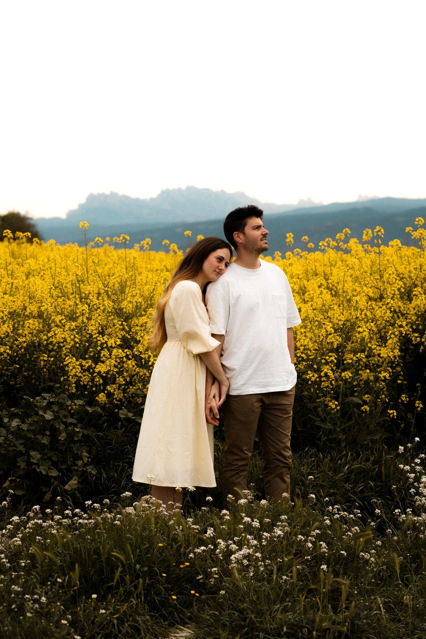 Una pareja de jóvenes en un campo de flores amarillas y blancas, con montañas al fondo, en un día nublado.