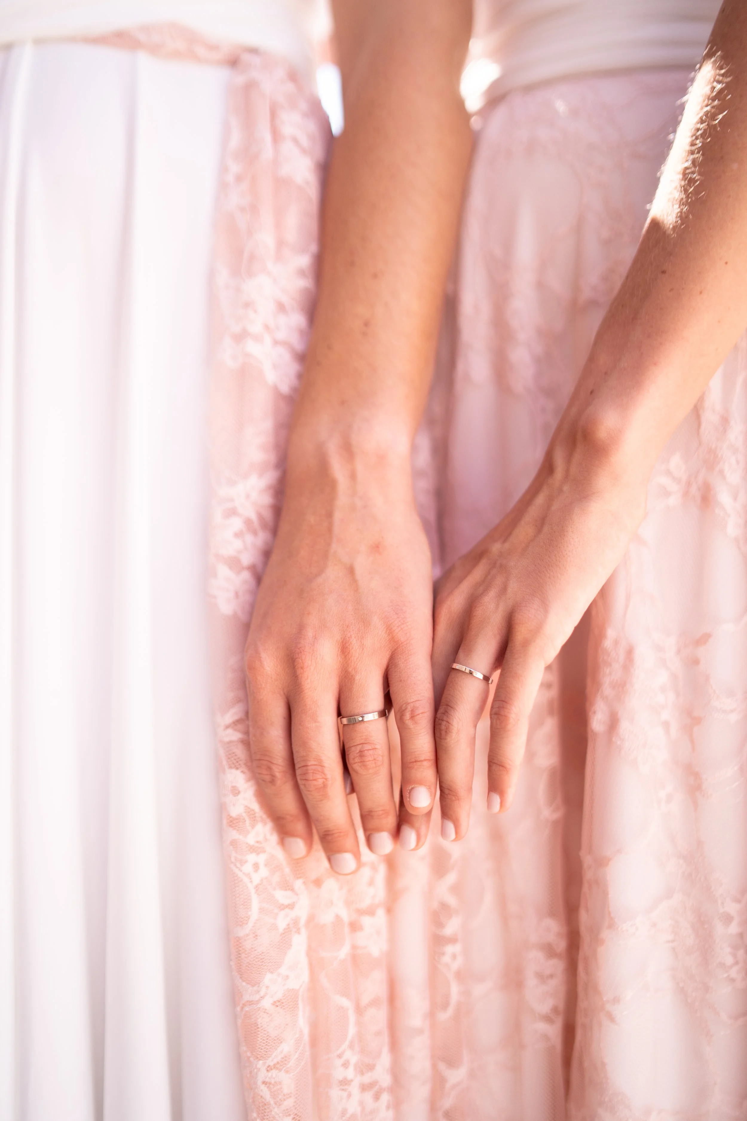 Dos manos de mujeres con anillos, vestidas con ropas de encaje y seda en tonos rosa y blanco, entrelazadas suavemente.