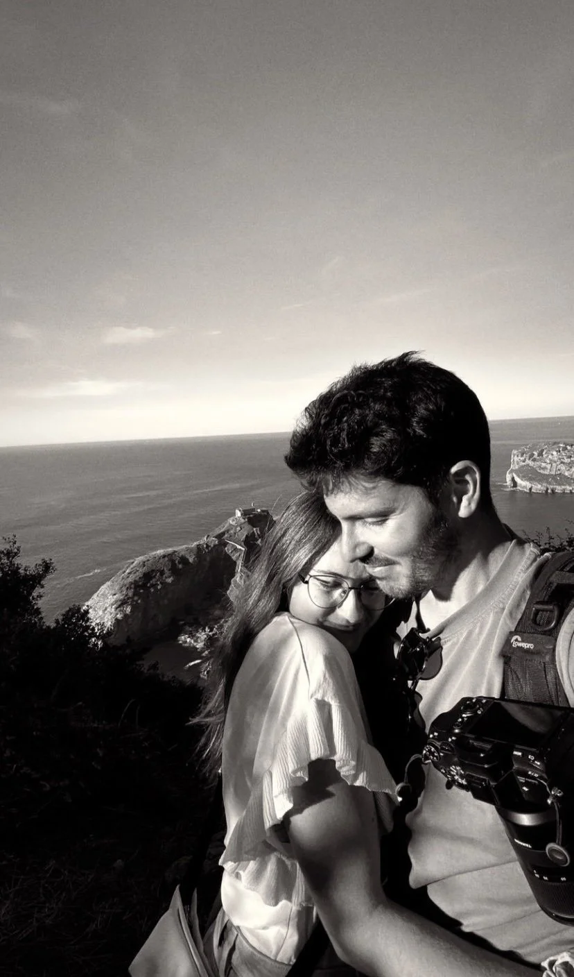 Dos personas abrazadas en un paisaje costero, vista del mar y acantilados, en una foto en blanco y negro.