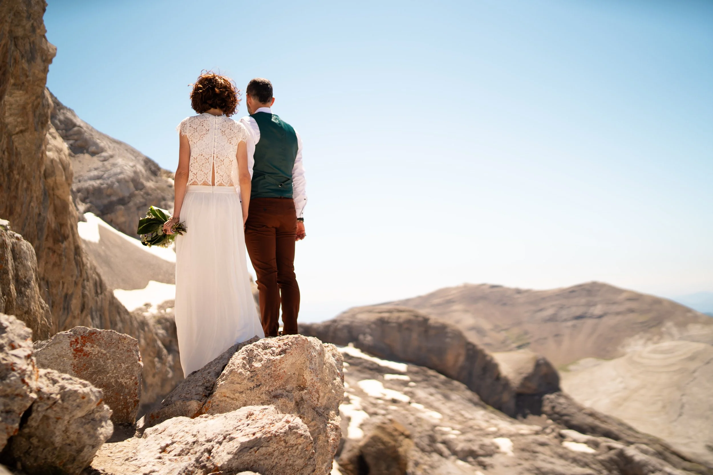 Pareja de boda de espaldas en un paisaje montañoso, ella lleva un vestido blanco y él lleva camisa blanca y chaleco verde, ella sostiene un ramo de flores.
