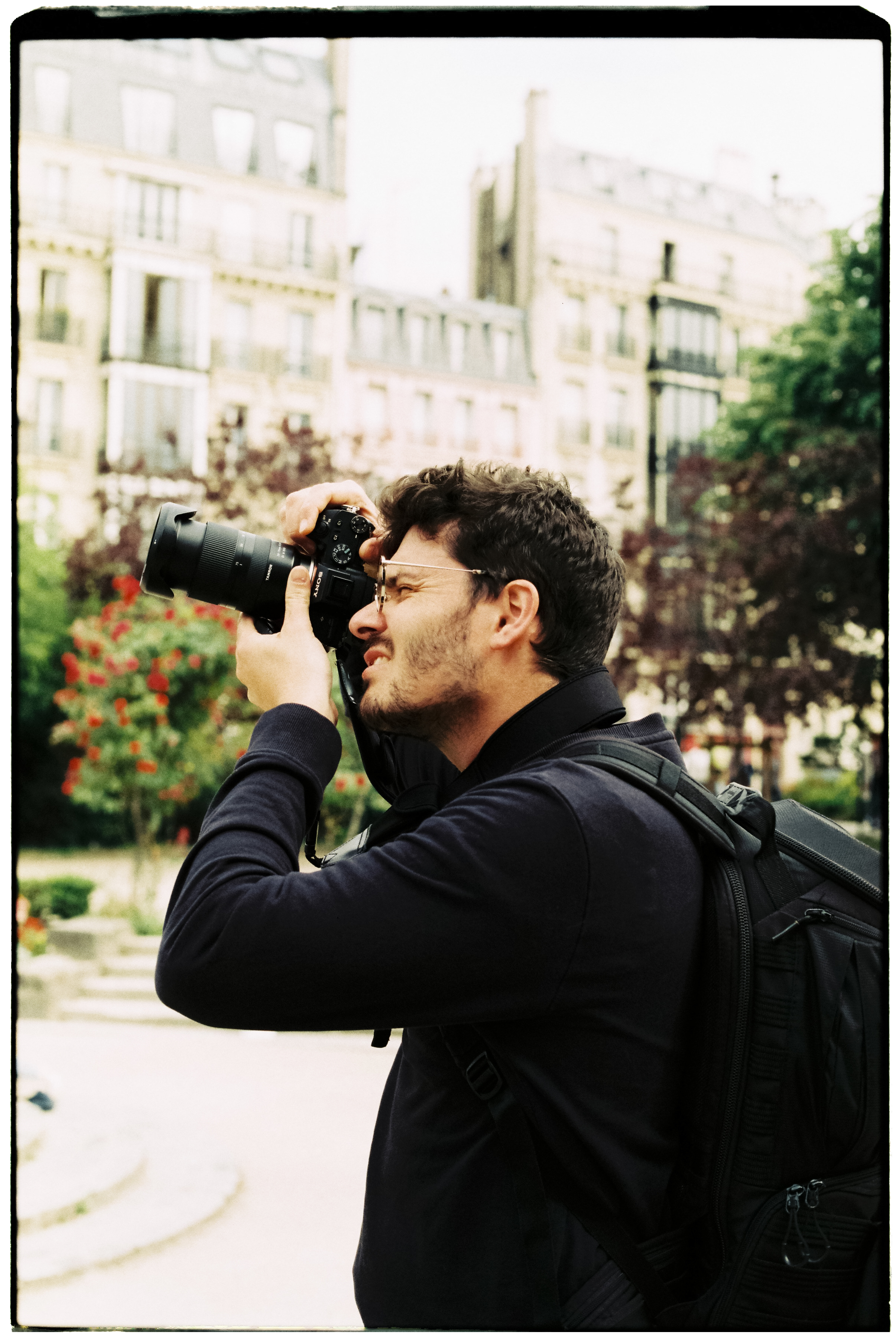Joven con mochila toma una fotografía con una cámara en un parque urbano con edificios y árboles de fondo.