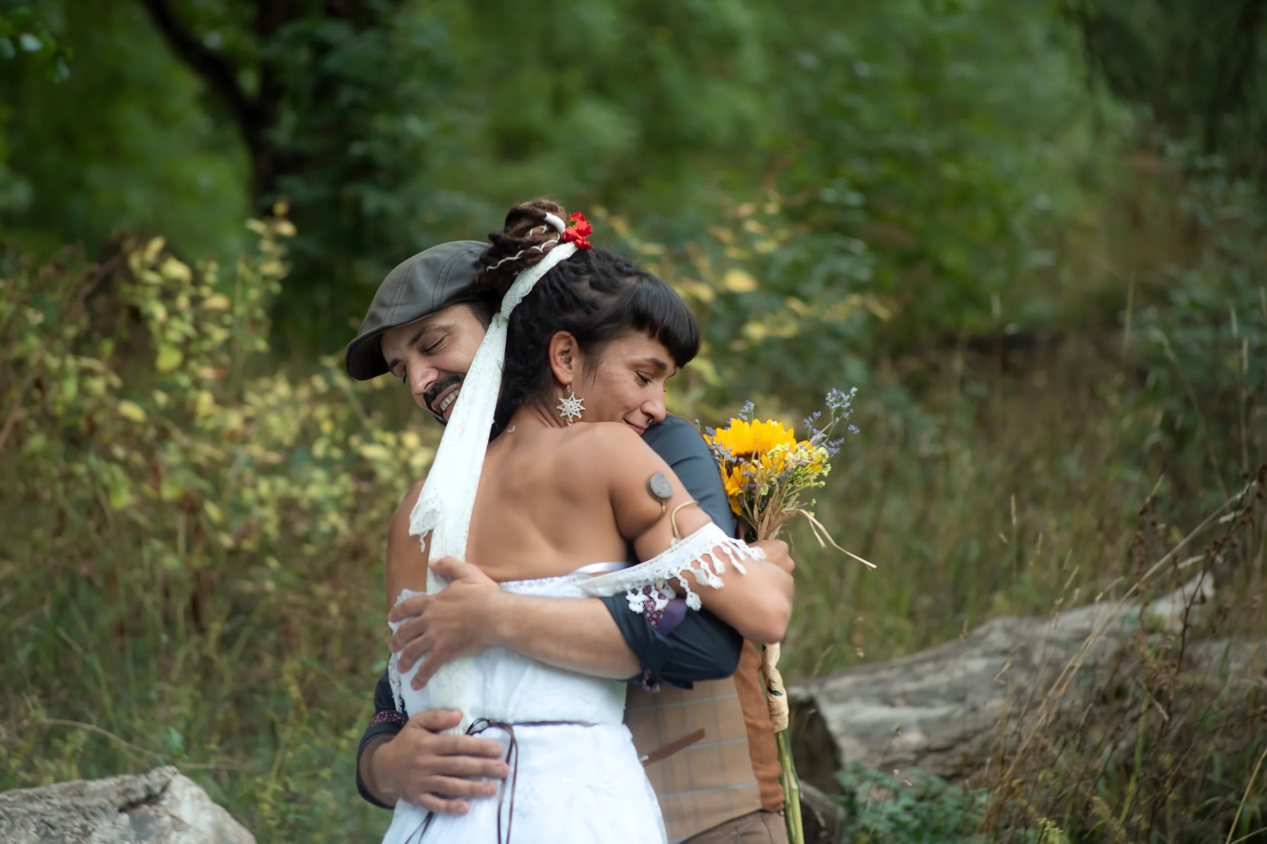 Una pareja feliz abrazándose en un entorno natural del bosque, con la mujer sosteniendo un ramo de flores.