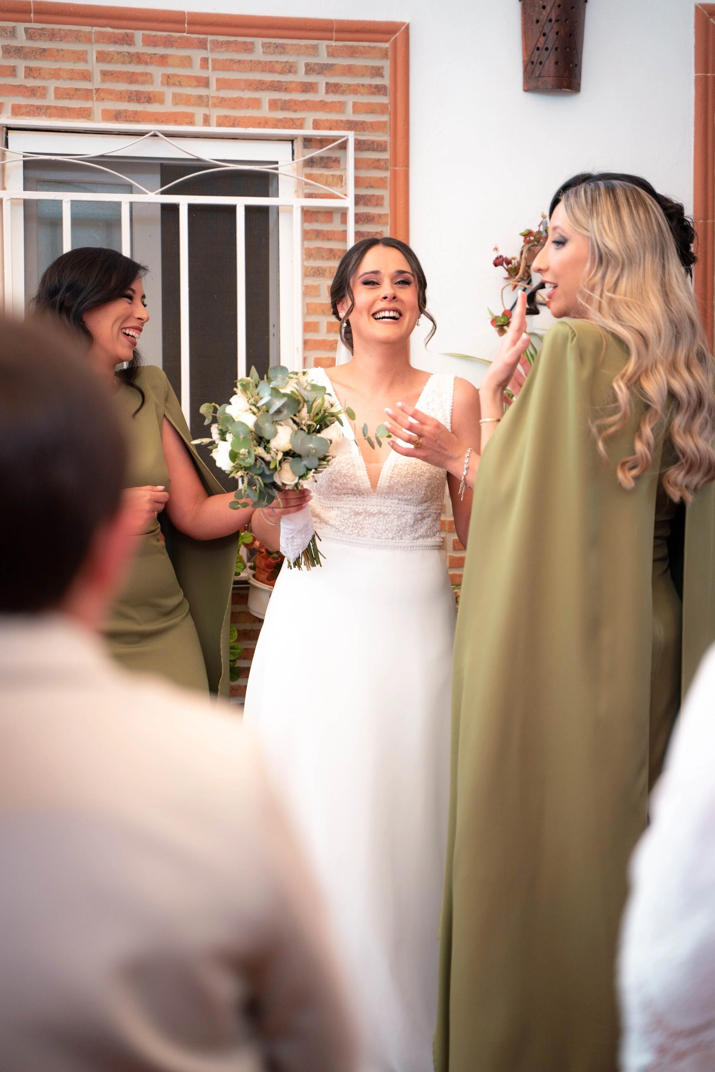 Mujer casada con vestido blanco social riendo y rodeada de tres mujeres que la felicitan, en un ambiente luminoso y decorado con paredes de ladrillo y plantas.