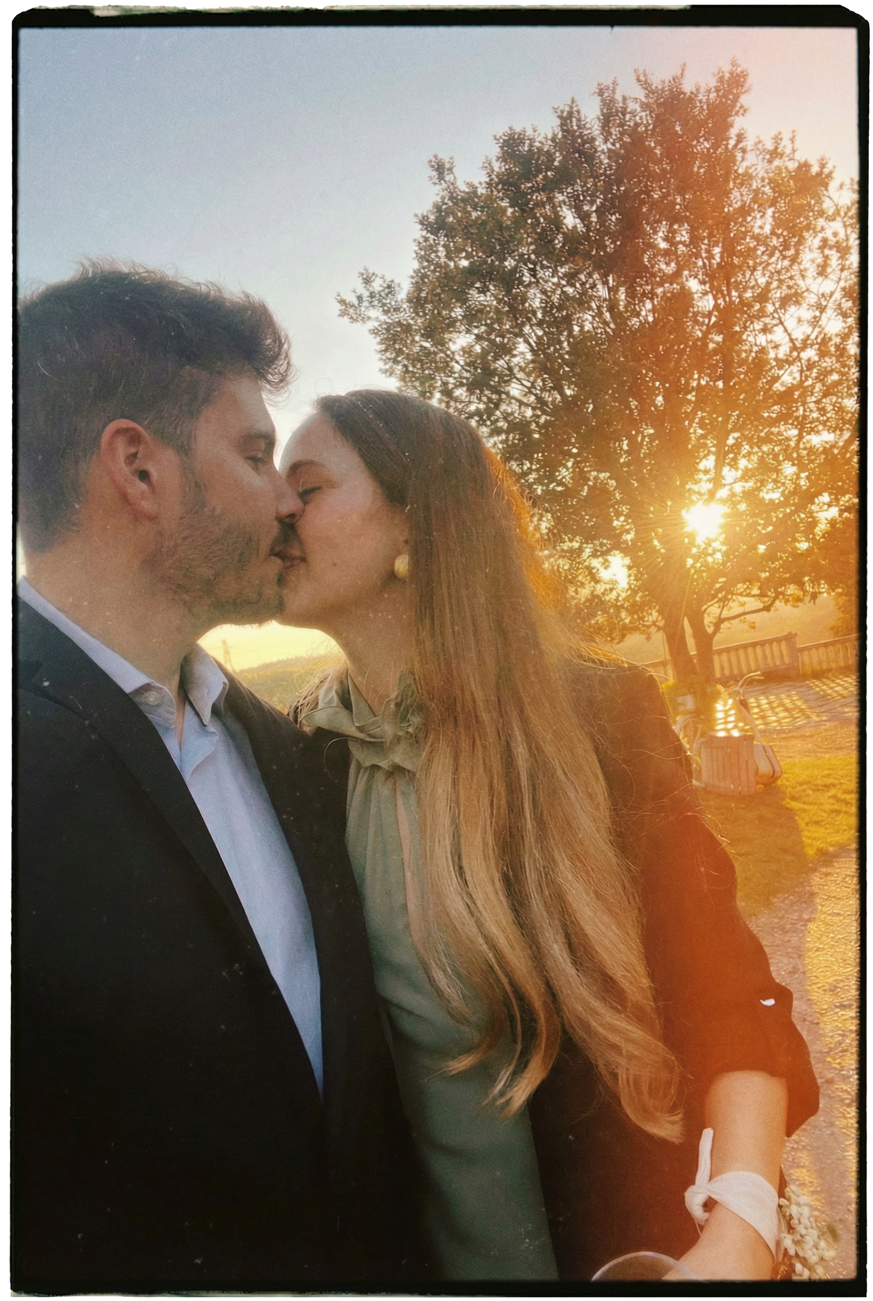 Una pareja enamorada se besa al atardecer en un parque, con un árbol grande y el sol poniéndose en el fondo.