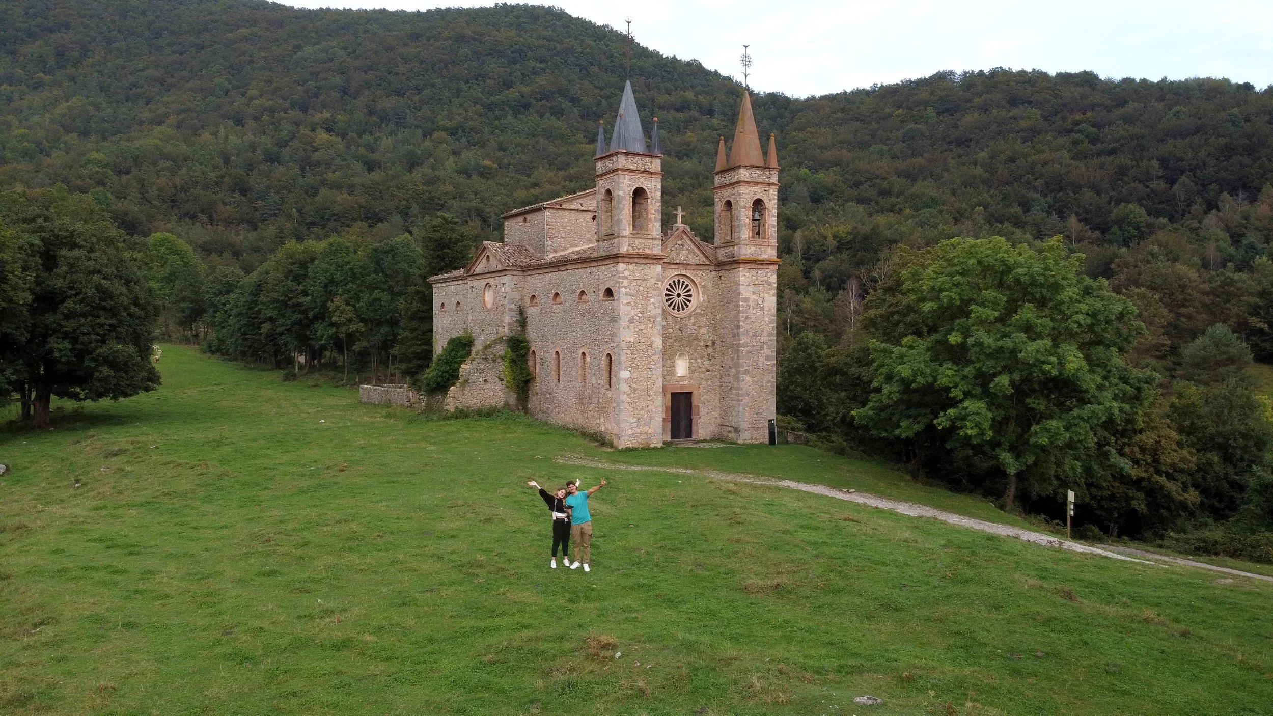 Una iglesia antigua de piedra en medio de un verde paisaje con árboles grandes y colinas de fondo, y dos personas celebrando en primer plano.