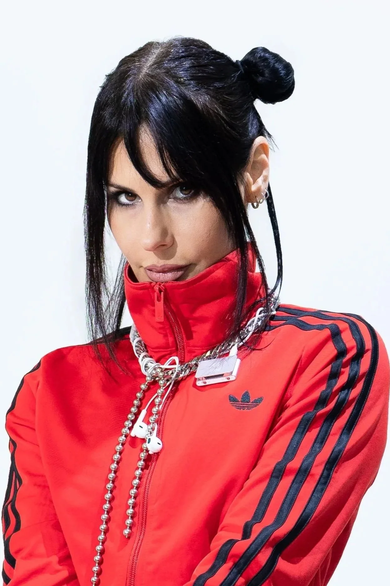 DJ REBEKAH.  who is a famous woman DJ, with dark hair styled in a bun, wearing a red Adidas jacket with black stripes and a gold chain necklace, with earbuds hanging from her neck.