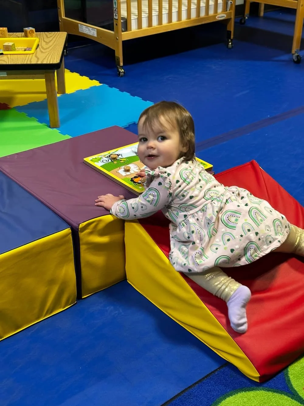 A young girl wearing a rainbow print dress plays on a soft play area with colorful padded blocks and a puzzle at Kid Champion daycare and early childhood education center (infants and toddlers) in Little Rock, Arkansas.
