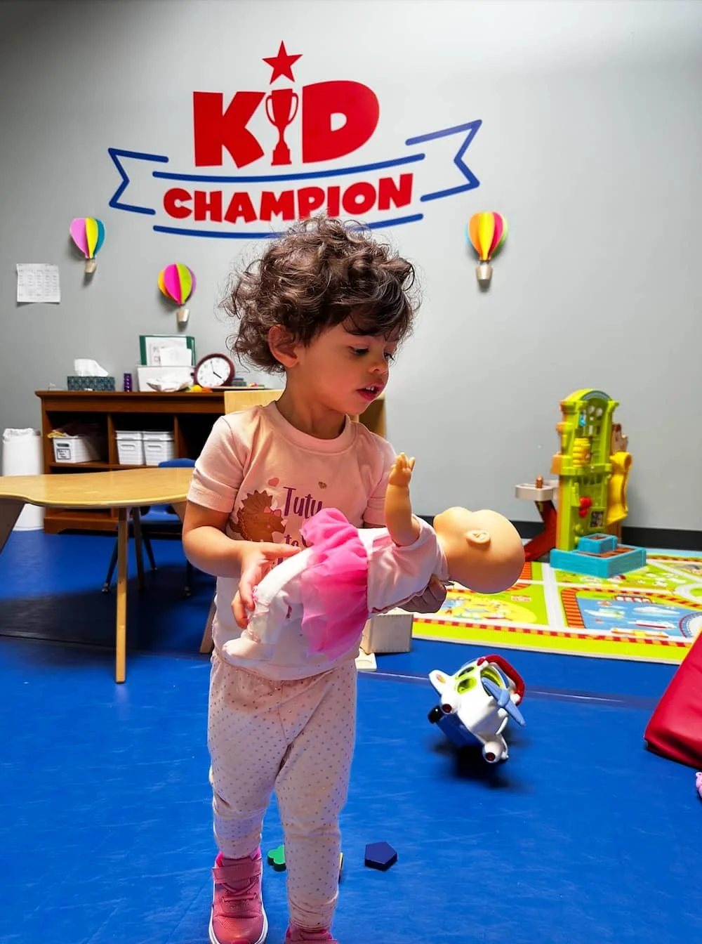 A young girl with curly hair playing with a baby doll in a colorful play area at a Kid Champion facility. The backdrop features a wall with the Kid Champion logo and various toys and decorations.