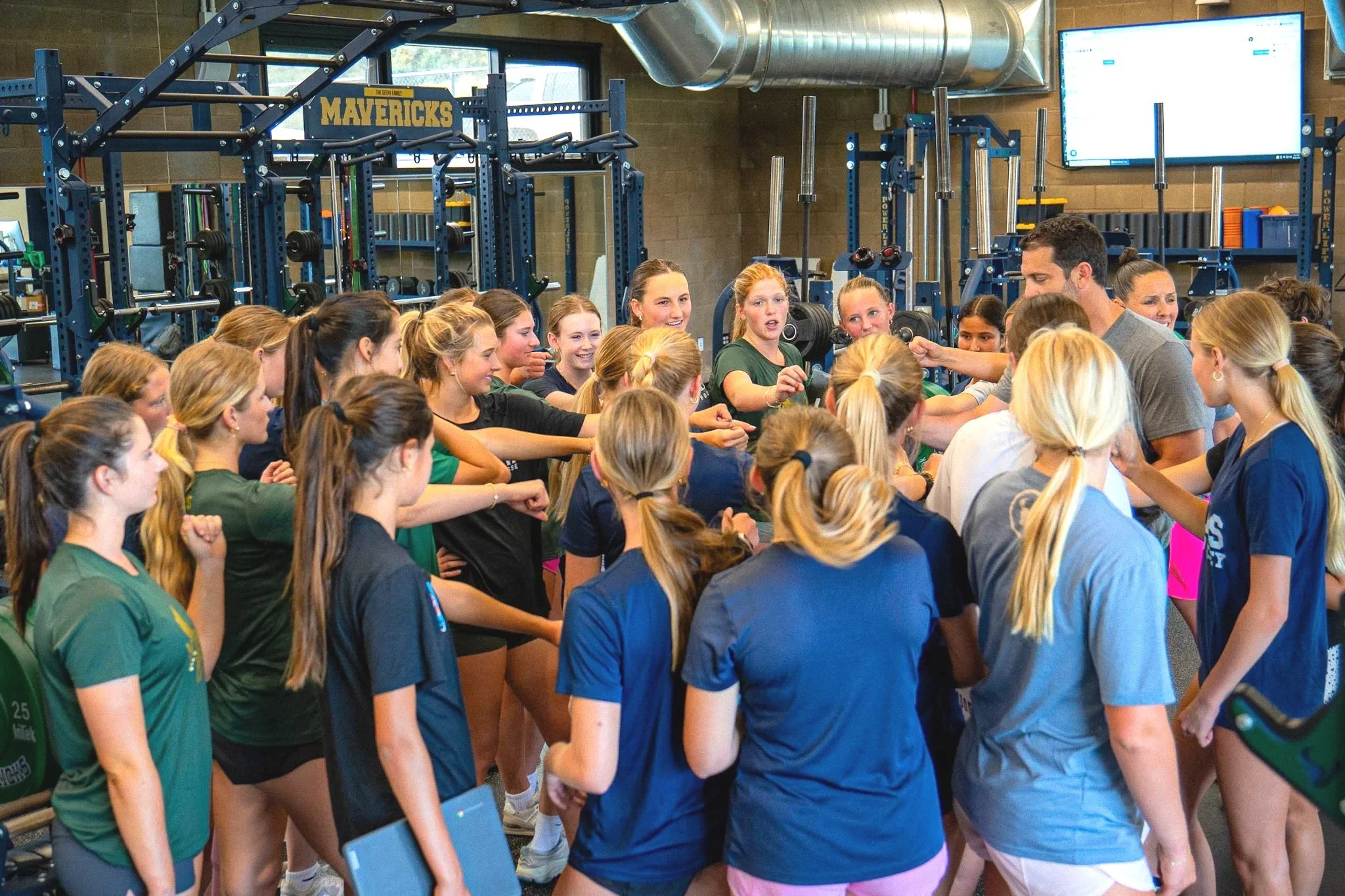 Women and girls gathered around a fitness trainer in a gym, listening and participating in a group workout session. The gym has weightlifting equipment and large windows.