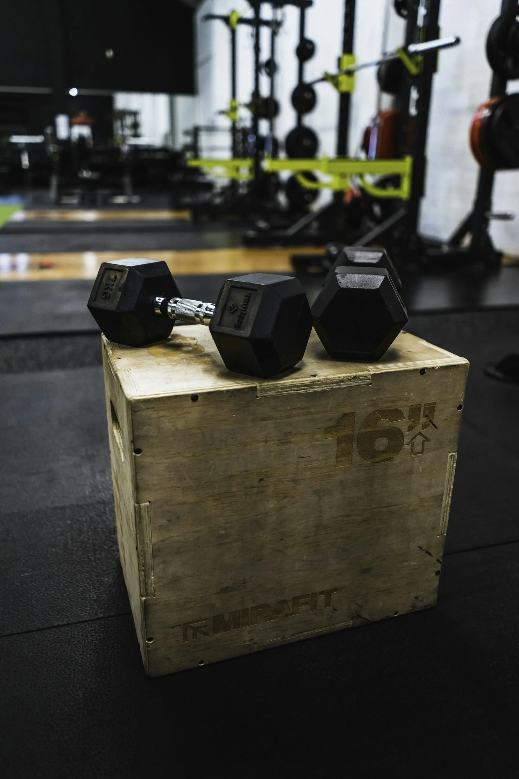 A pair of black dumbbells resting on a wooden plyo box in a gym with weightlifting equipment in the background.