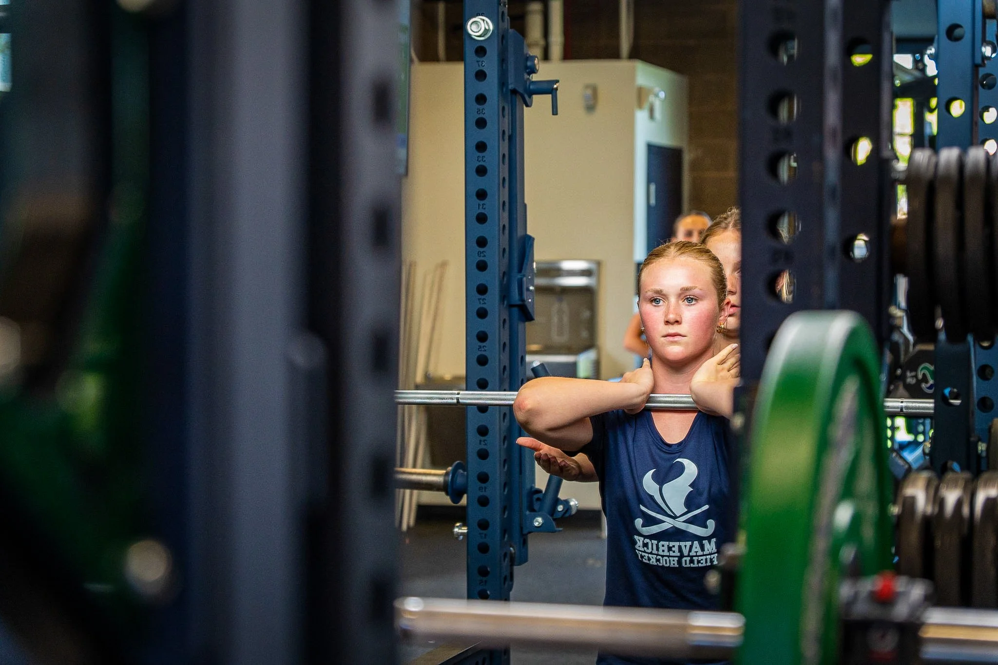 A young girl in a gym lifting a barbell, framed by gym equipment and weights.