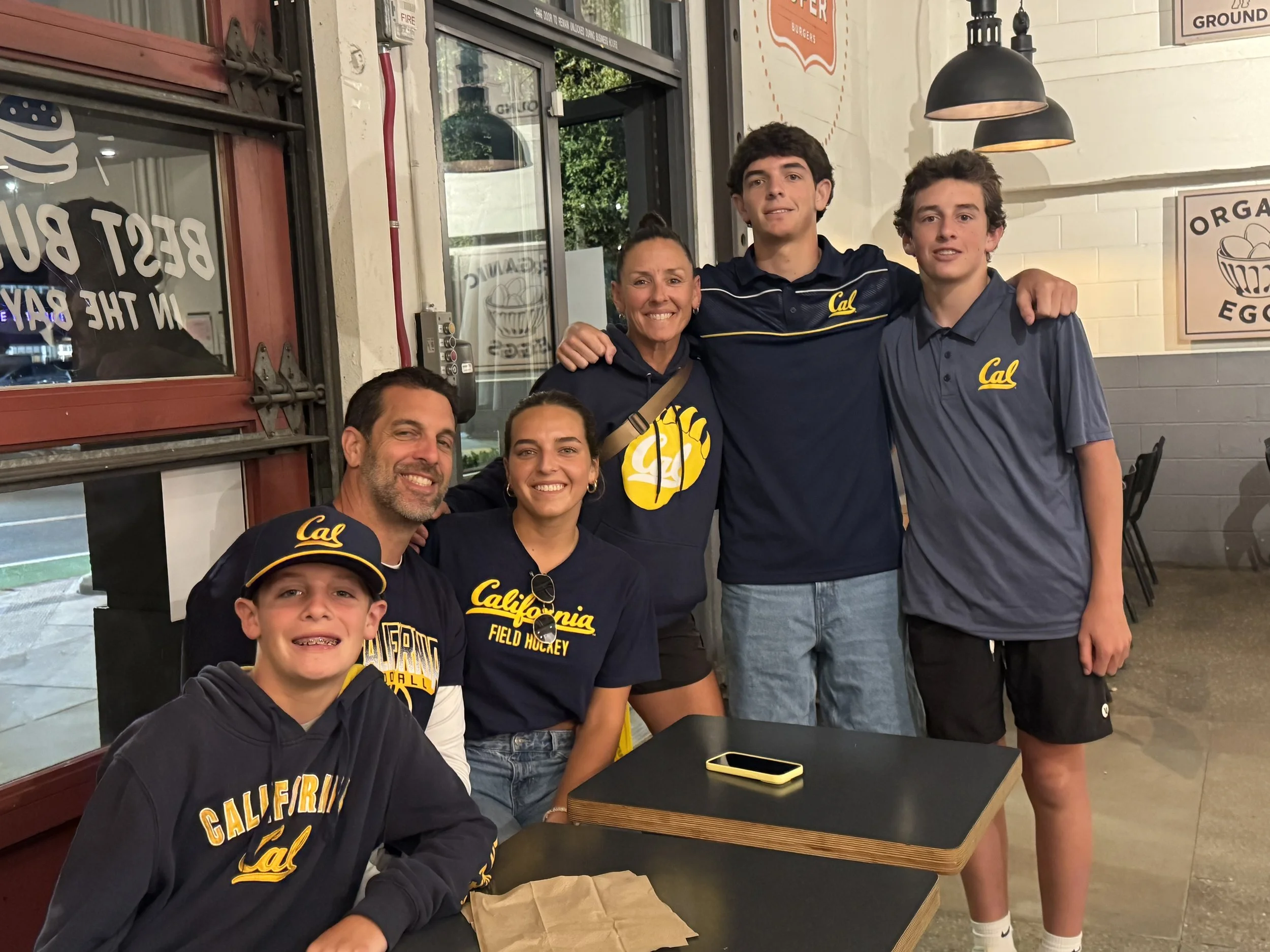 A group of five people, four males and one female, posing together inside a restaurant with some signs on the wall and taking a selfie. Most of them are wearing navy blue shirts or hoodies with the word 'Cal' and 'California' on them. They appear to be happy, smiling, and enjoying their time together.